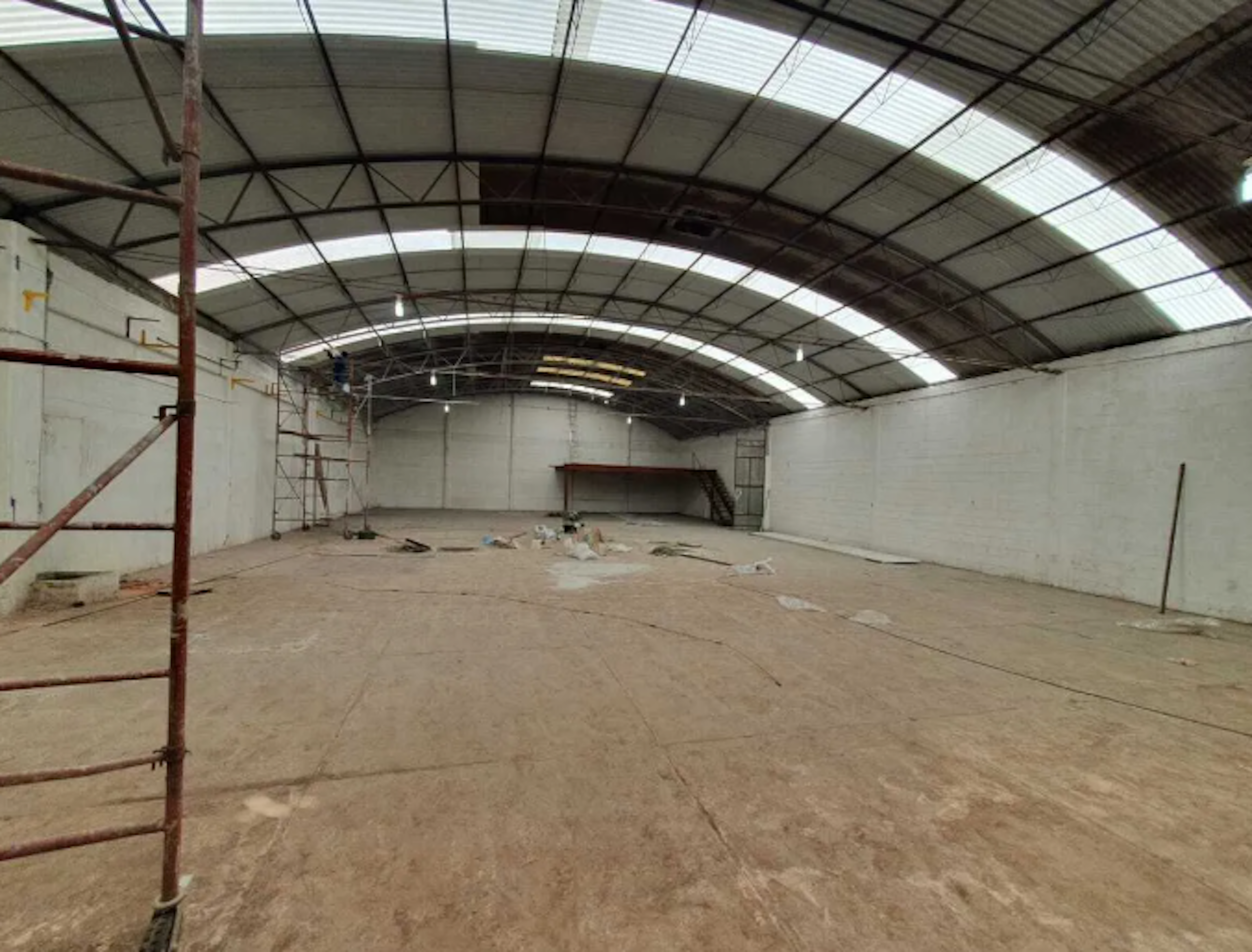 La imagen muestra el interior de una bodega industrial en proceso de remodelación. Se observa un amplio espacio con techo abovedado de metal y paredes blancas. El piso está cubierto de escombros y materiales de construcción. Hay andamios y una escalera visible, indicando trabajos en curso. La iluminación natural proviene de paneles translúcidos en el techo. El espacio parece estar en las etapas iniciales de una renovación o readaptación.