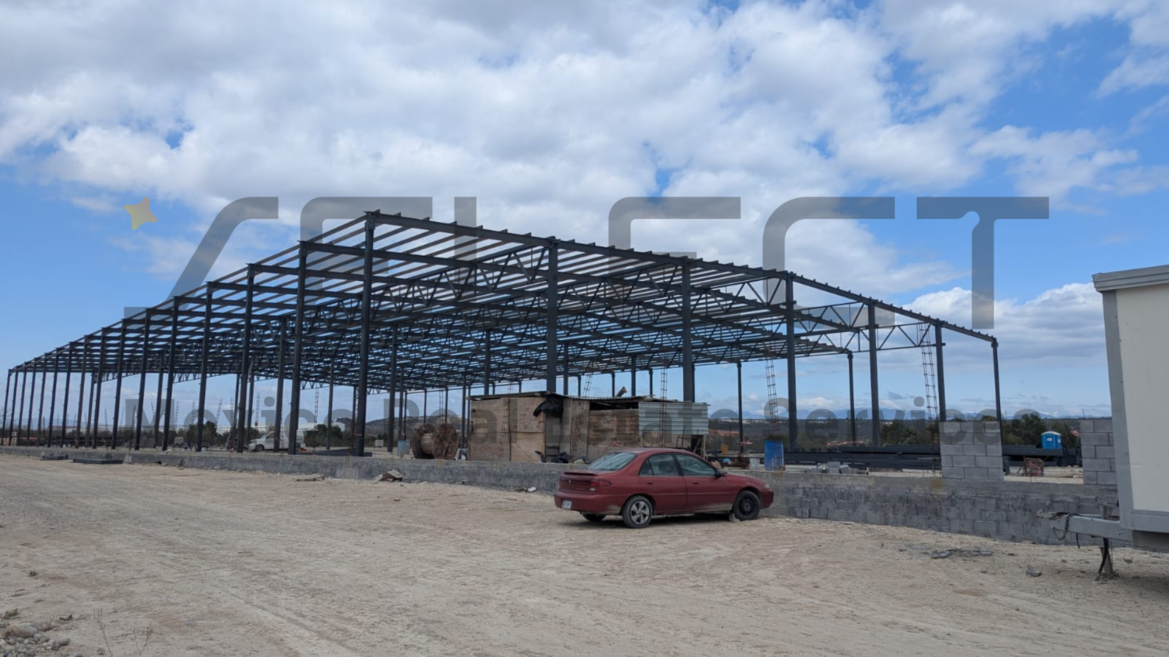 La imagen muestra una estructura de acero en construcción, probablemente destinada a ser un gran almacén o nave industrial. El esqueleto metálico del edificio está completo, pero aún sin paredes ni techo. En primer plano se ve un terreno sin pavimentar y un automóvil rojo estacionado. El cielo es azul con algunas nubes, sugiriendo un día despejado en una zona de desarrollo industrial o comercial.