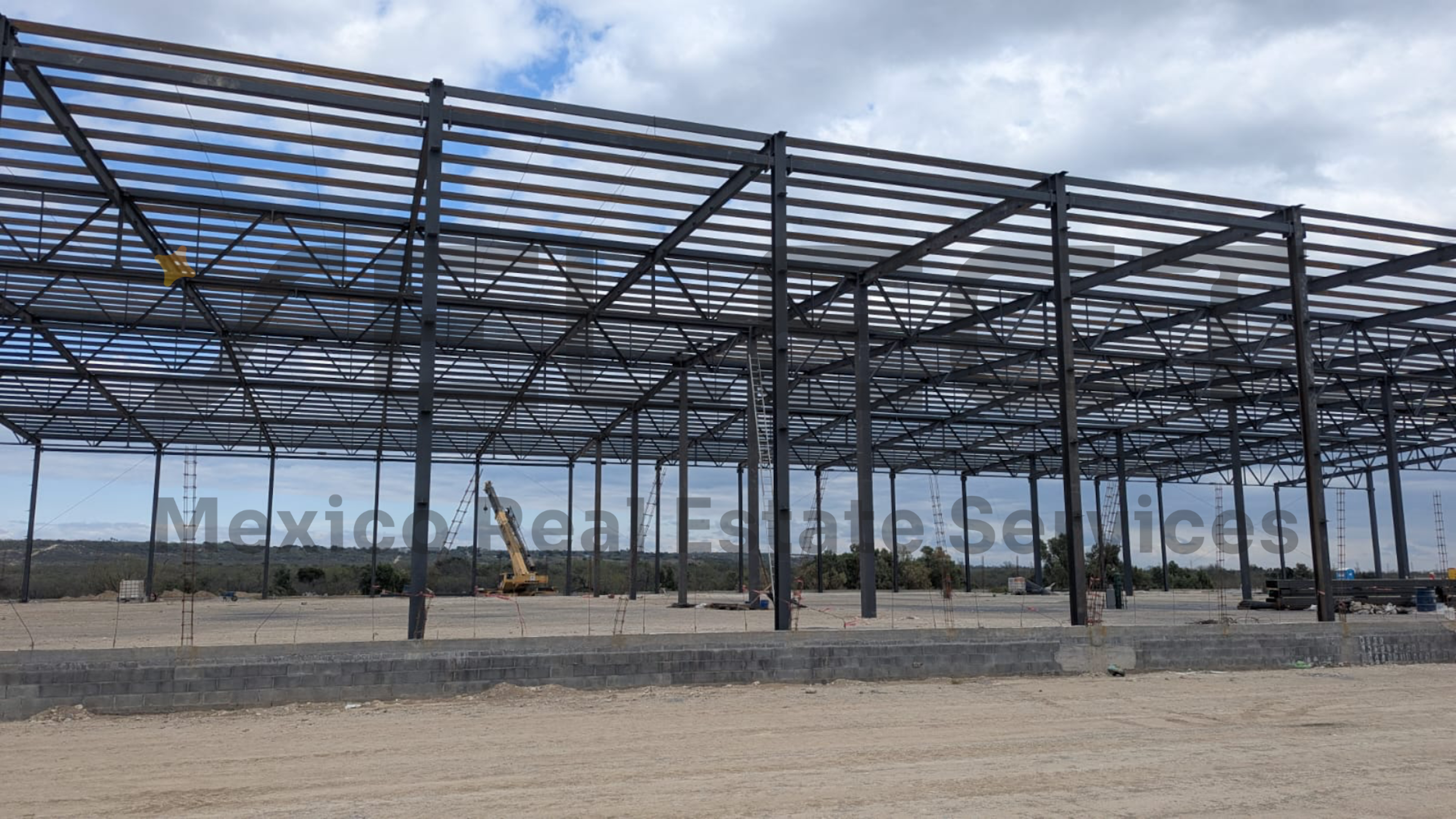 La imagen muestra una estructura de acero en construcción, probablemente para un almacén o nave industrial. Se observa un amplio marco metálico con vigas y soportes, típico de edificaciones comerciales o industriales. El sitio parece estar en una zona desértica o rural, con un paisaje abierto al fondo. La presencia de maquinaria pesada sugiere que el proyecto está en pleno desarrollo. El texto superpuesto Mexico Real Estate Services indica que esto podría ser parte de un desarrollo inmobiliario en México.