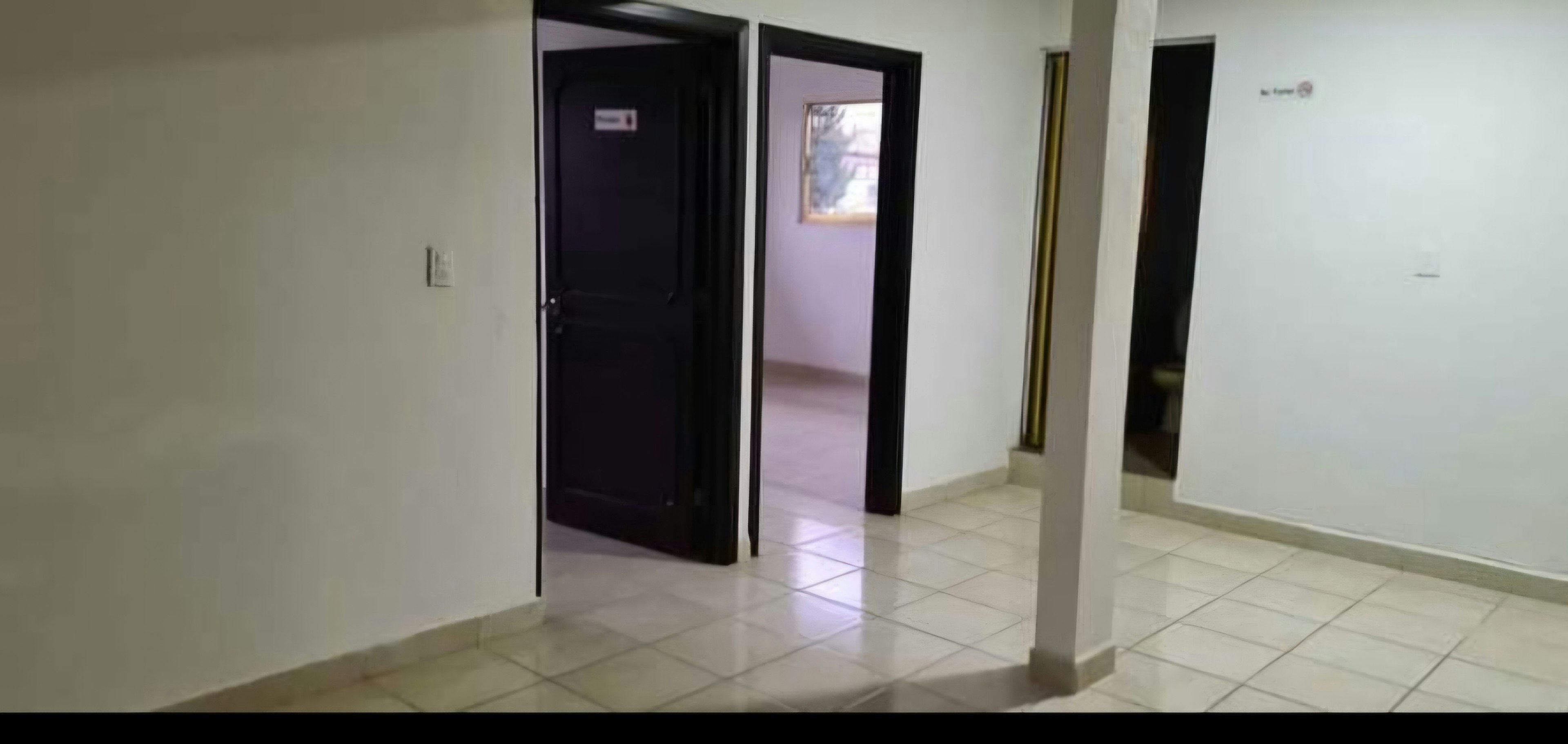 The image shows an interior hallway of a property, likely an apartment or office space. It features white tiled flooring, white walls, and dark wooden doors. One door is open, revealing a glimpse of another room with a window. The space appears clean and well-maintained, with a modern, minimalist aesthetic. The contrast between the dark doors and light walls creates a sleek, contemporary look.