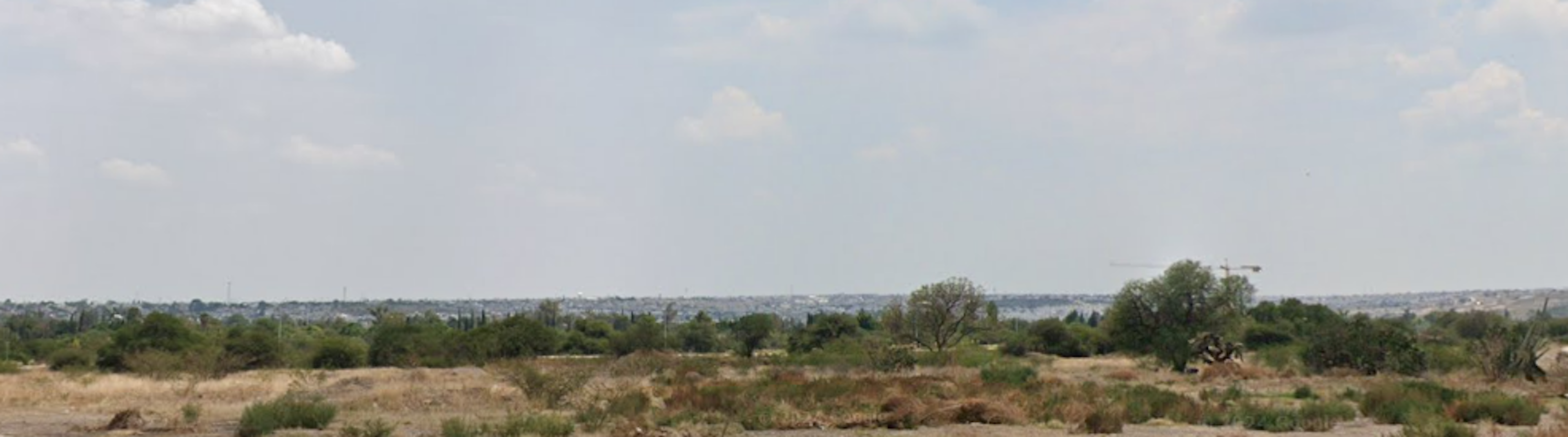 La imagen muestra un terreno extenso y plano en una zona semiárida. Se observa vegetación baja y dispersa típica de climas secos. En el horizonte se aprecia una ciudad o zona urbana, lo que sugiere que este terreno podría ser una oportunidad para desarrollo inmobiliario en las afueras de una zona poblada. El cielo despejado y la vista panorámica indican potencial para proyectos residenciales o comerciales con buenas vistas.