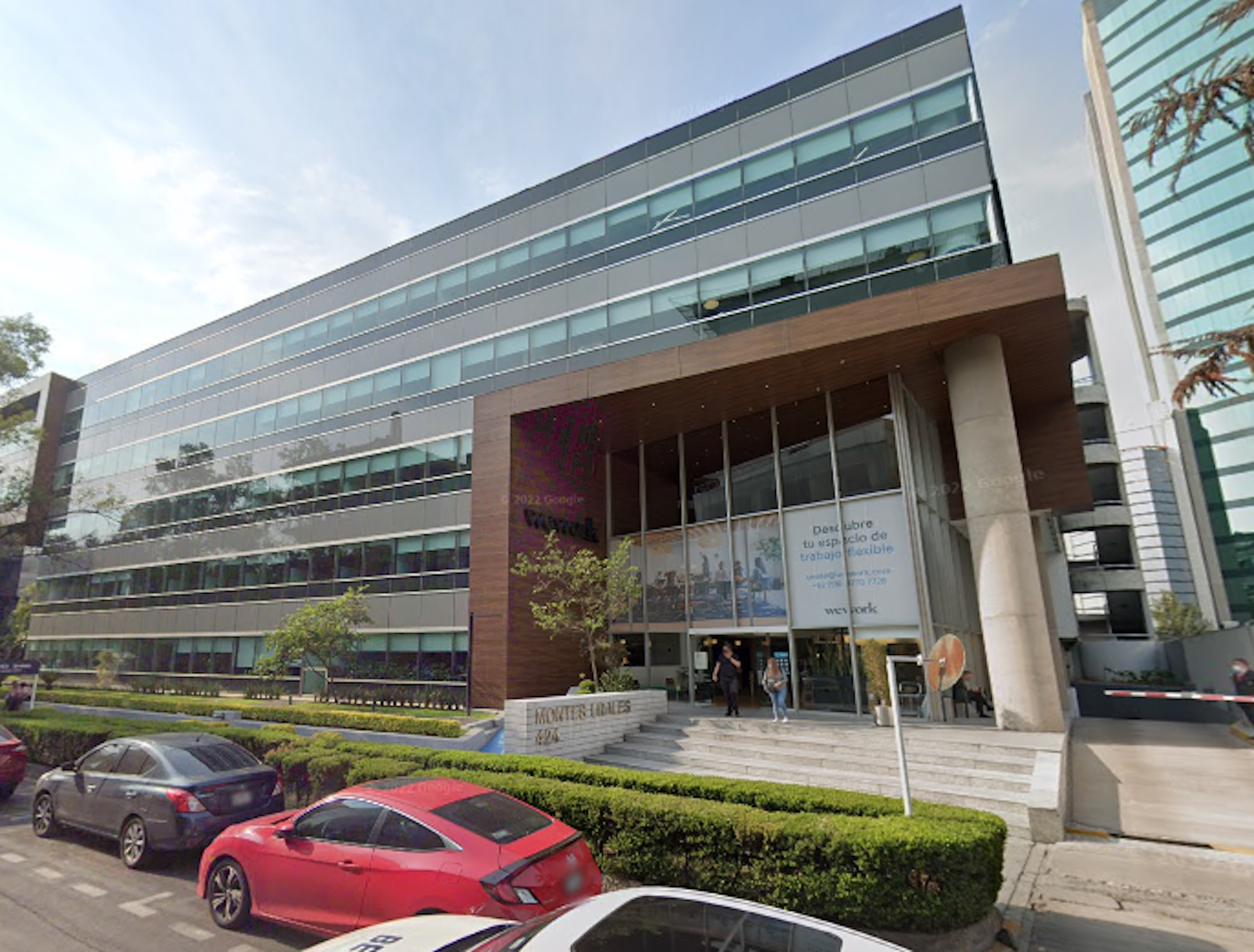 La imagen muestra un moderno edificio de oficinas con fachada de vidrio y acero. Tiene una entrada principal con detalles en madera y un letrero de WeWork. El edificio está rodeado de áreas verdes bien cuidadas y hay algunos autos estacionados frente a él. La arquitectura es contemporánea y el edificio parece ser parte de un complejo empresarial o zona financiera.