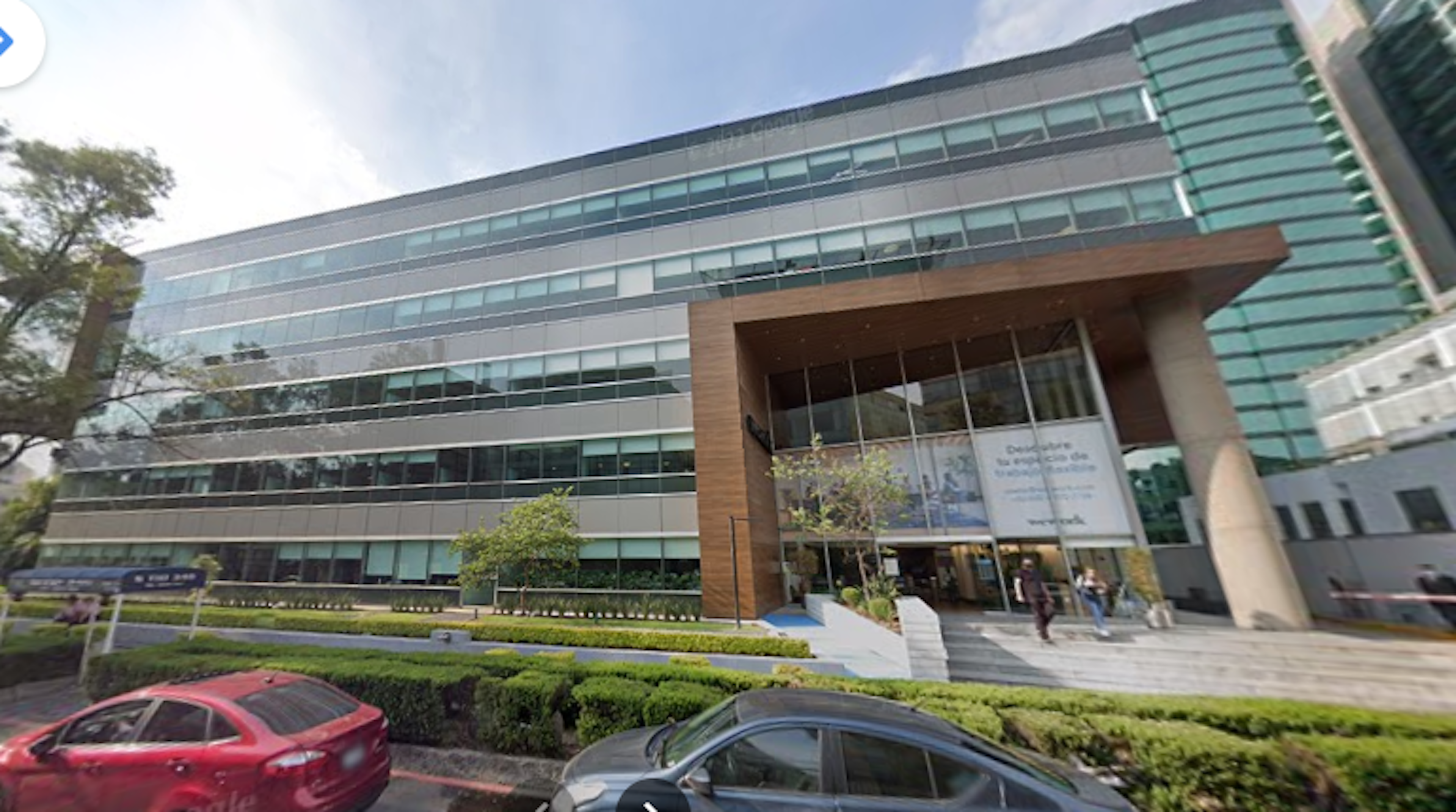 La imagen muestra un edificio de oficinas moderno con fachada de vidrio y acero. Tiene múltiples pisos y un diseño contemporáneo con una entrada prominente adornada con madera. El edificio está rodeado de áreas verdes y un estacionamiento en el frente. La arquitectura sugiere que es un espacio comercial de alta gama en una zona urbana desarrollada, probablemente utilizado para oficinas corporativas o servicios profesionales.