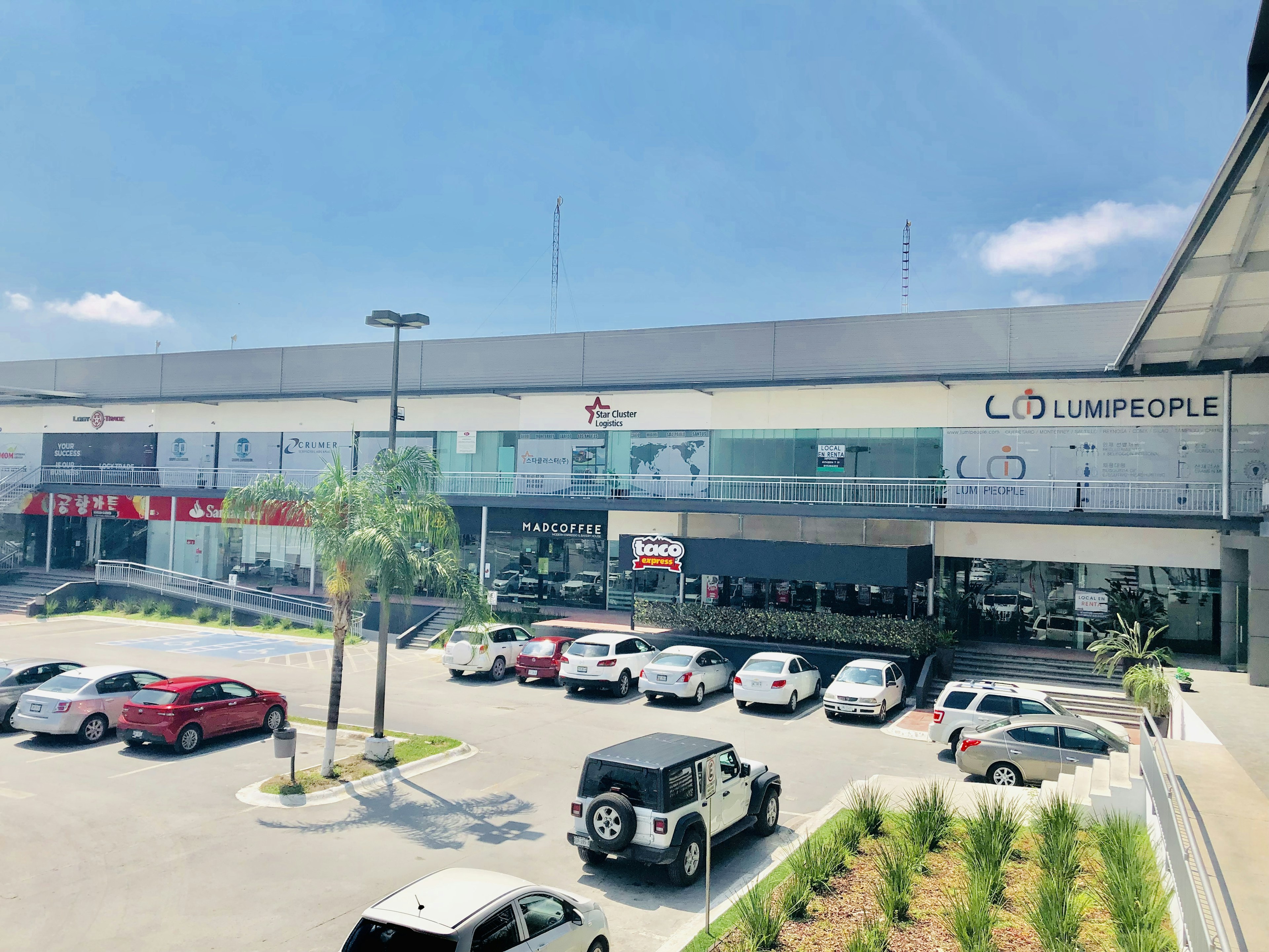 La imagen muestra un centro comercial moderno de dos niveles con estacionamiento al frente. El edificio tiene fachada blanca con logos de negocios como Lumipeople y Madcoffee. Hay una variedad de vehículos estacionados, incluyendo un Jeep. Se observa vegetación tropical como palmeras. El cielo es azul con algunas nubes, sugiriendo un clima agradable. La propiedad parece estar en buenas condiciones y bien mantenida.