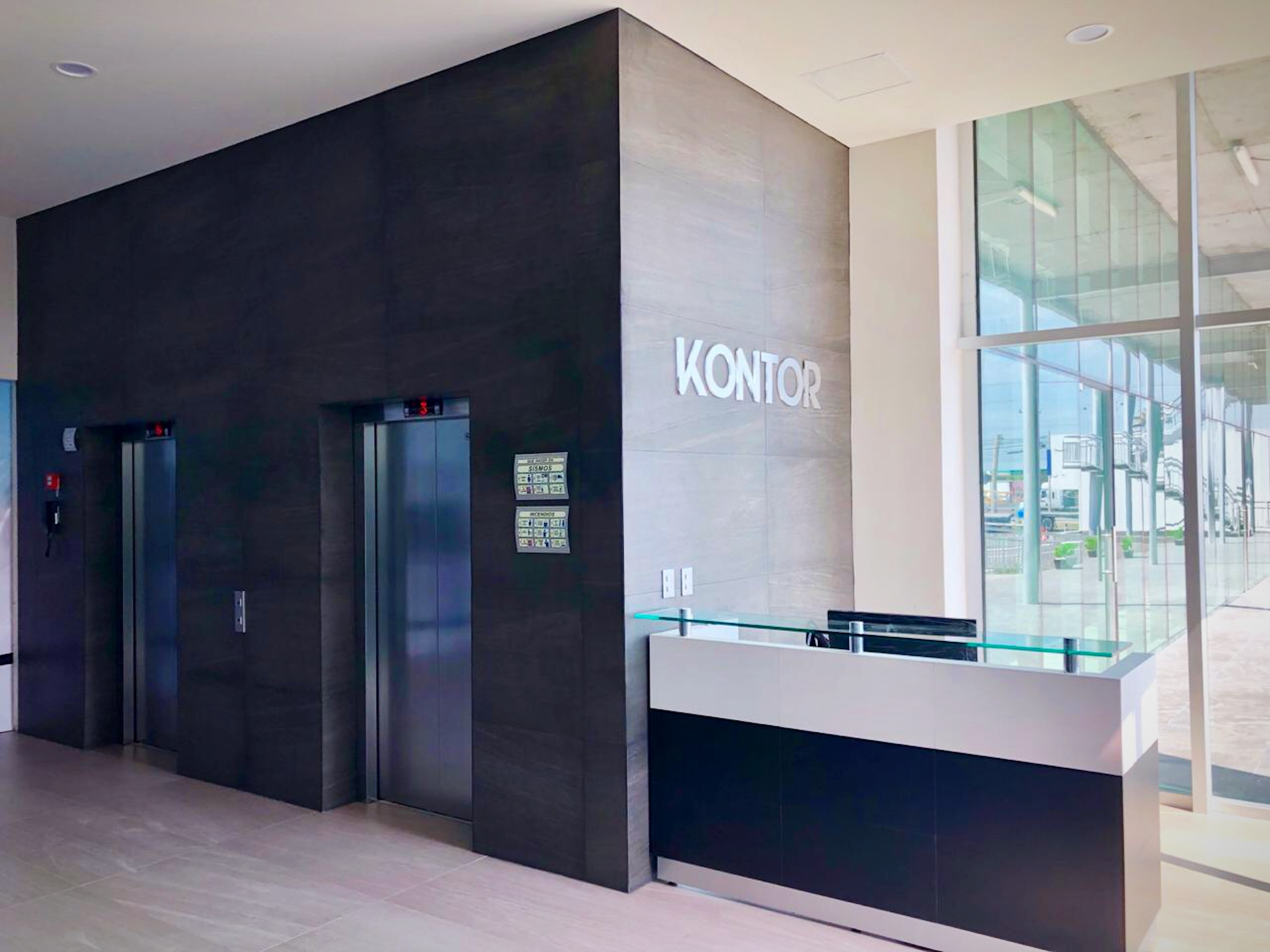 La imagen muestra el interior de un edificio de oficinas moderno. Se ve un área de recepción con el letrero KONTOR en la pared. Hay dos ascensores con puertas de acero inoxidable y un mostrador de recepción blanco y negro con detalles en vidrio. El diseño es minimalista, con paredes oscuras contrastando con áreas más claras y grandes ventanales que permiten la entrada de luz natural.