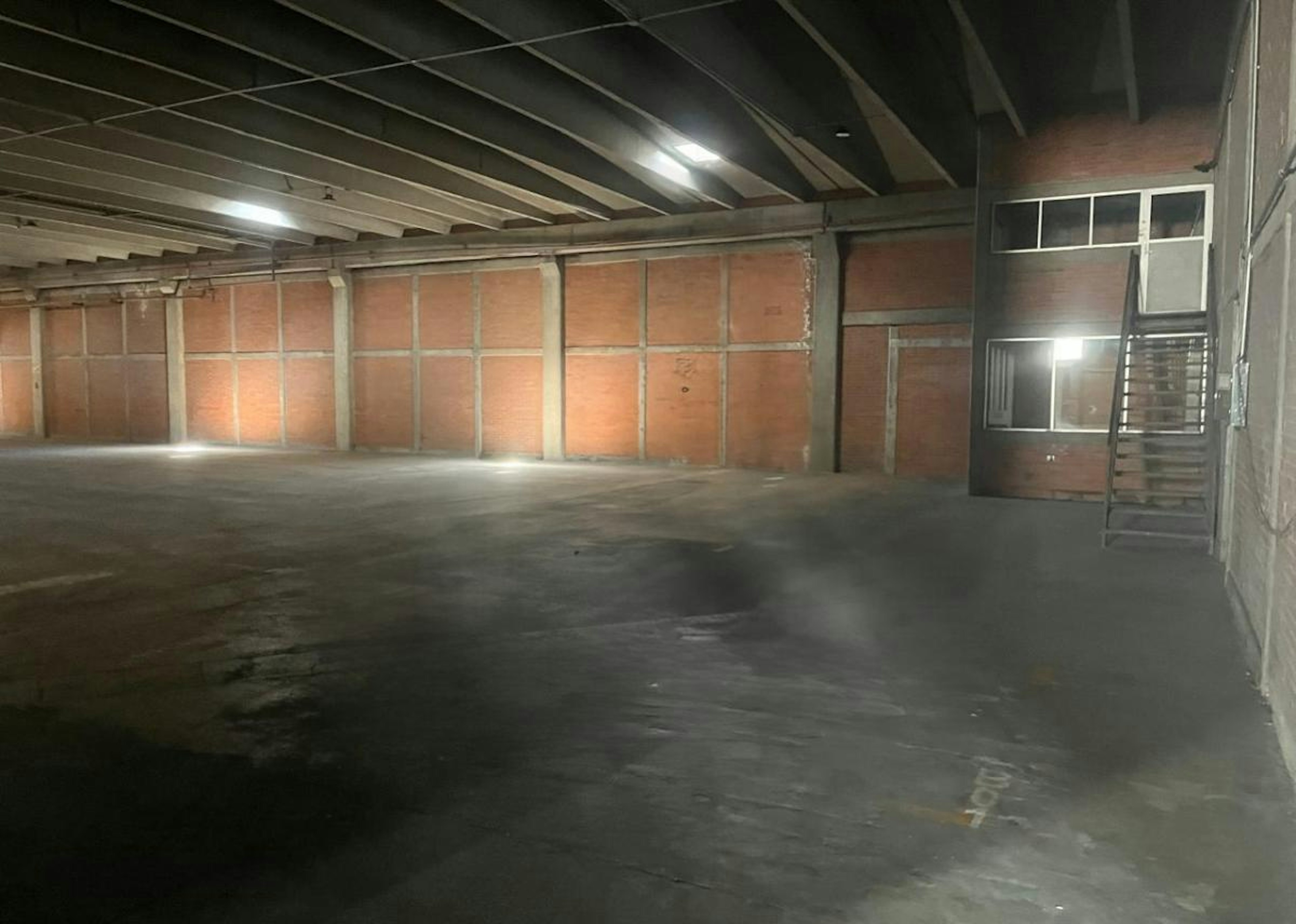 La imagen muestra un amplio espacio industrial vacío con paredes de ladrillo, techo con vigas expuestas y piso de concreto. Se ve una pequeña oficina o área cerrada en el fondo. El espacio parece estar en proceso de remodelación o preparación para uso logístico. Hay un logo superpuesto de SIL - Strategic Industry Logistics que sugiere el propósito del lugar para operaciones industriales y de logística.