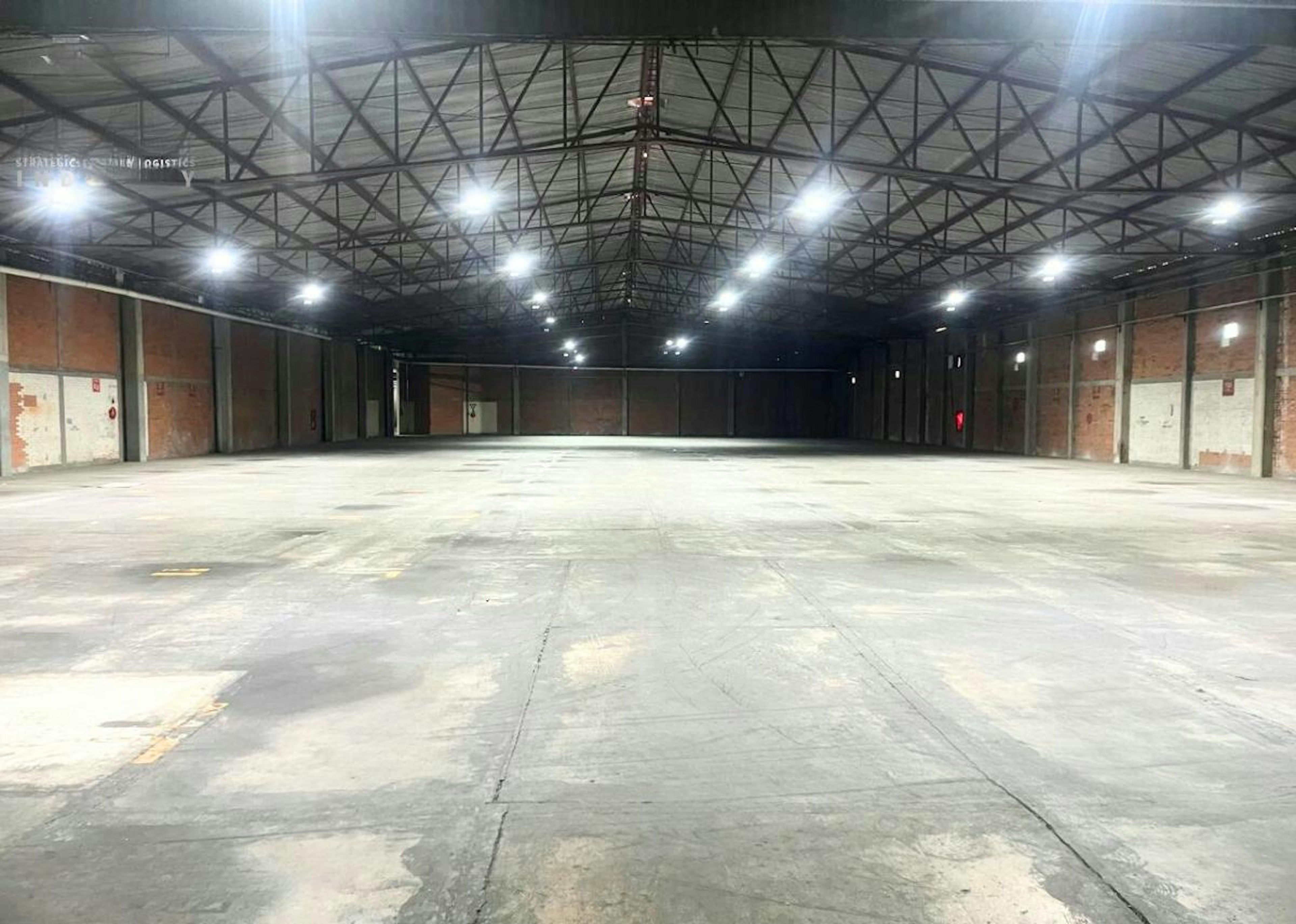 La imagen muestra un amplio almacén industrial vacío con techo alto de estructura metálica y múltiples luces. El piso es de concreto y las paredes laterales son de ladrillo. El espacio está completamente despejado, ideal para almacenamiento o distribución. Se observa el logotipo de Strategic Industry Logistics y un número de teléfono de contacto, sugiriendo que el espacio está disponible para alquiler o venta.