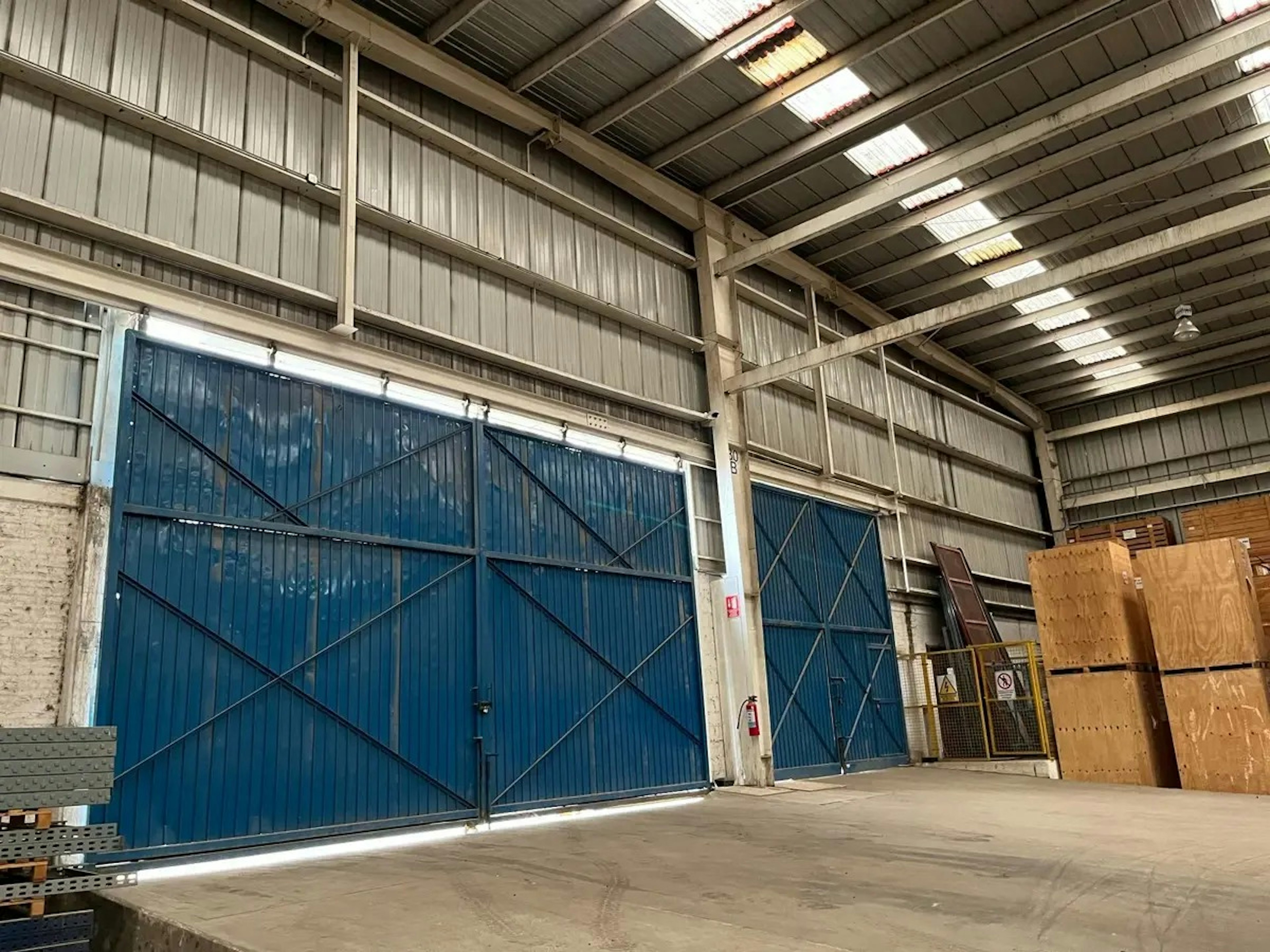 Esta imagen muestra el interior de un gran almacén o nave industrial. Se observan grandes puertas correderas azules con estructuras metálicas. El techo es alto con vigas expuestas y paneles translúcidos para iluminación natural. Las paredes son de metal corrugado. Se pueden ver algunos materiales de construcción y cajas almacenadas. El espacio parece amplio y funcional, ideal para almacenamiento o actividades industriales.