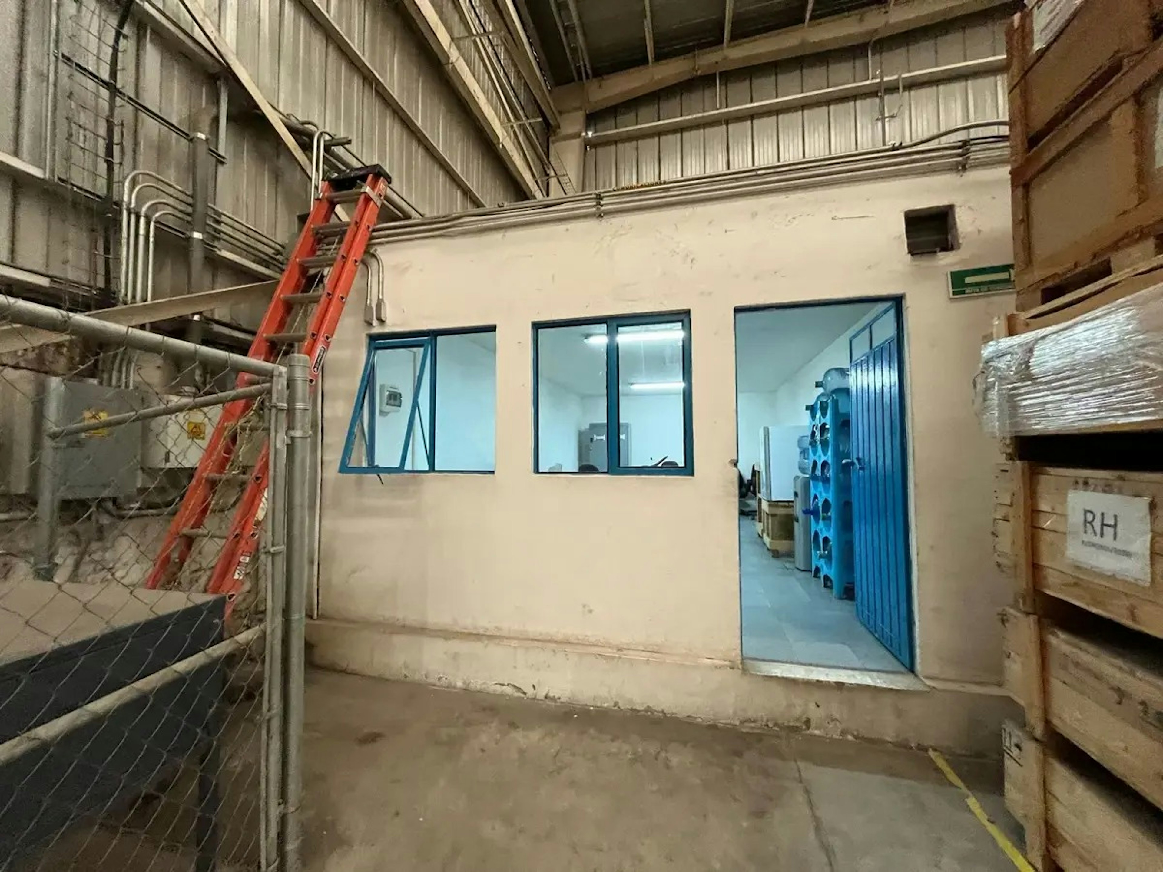 La imagen muestra el interior de un espacio industrial en proceso de renovación. Se observa una pared con ventanas y una puerta azul que da acceso a un área más iluminada. Hay una escalera roja apoyada contra la pared, tuberías expuestas en el techo y cajas almacenadas. El espacio parece estar en transición, posiblemente convirtiéndose en una oficina o área de almacenamiento más moderna dentro de un edificio industrial.