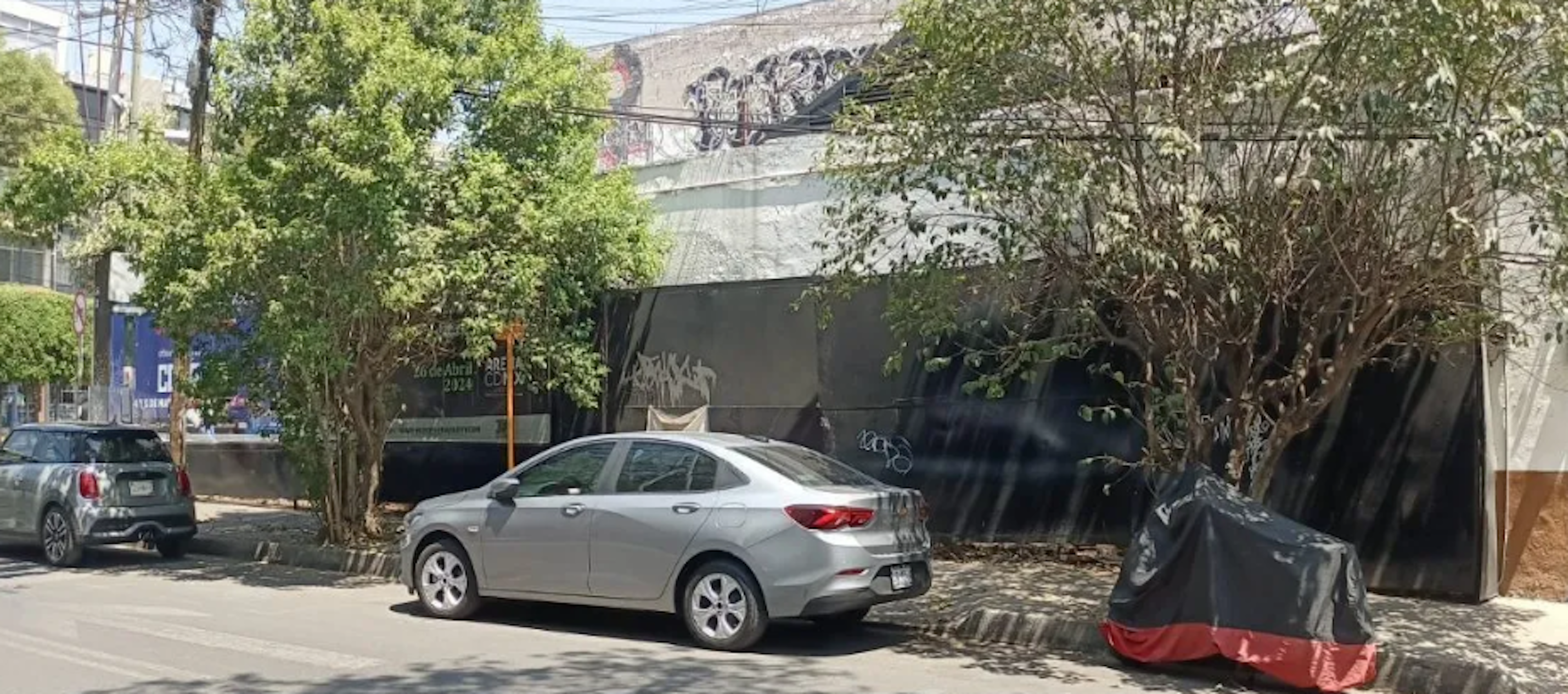 La imagen muestra una calle urbana con edificios bajos y árboles. Hay varios vehículos estacionados, incluyendo un sedán plateado en primer plano. Los edificios tienen fachadas grises y algunas paredes con grafitis. La vegetación proporciona sombra y un aspecto verde a la escena. El entorno sugiere una zona residencial o comercial de baja densidad, típica de áreas urbanas en desarrollo o en proceso de renovación.