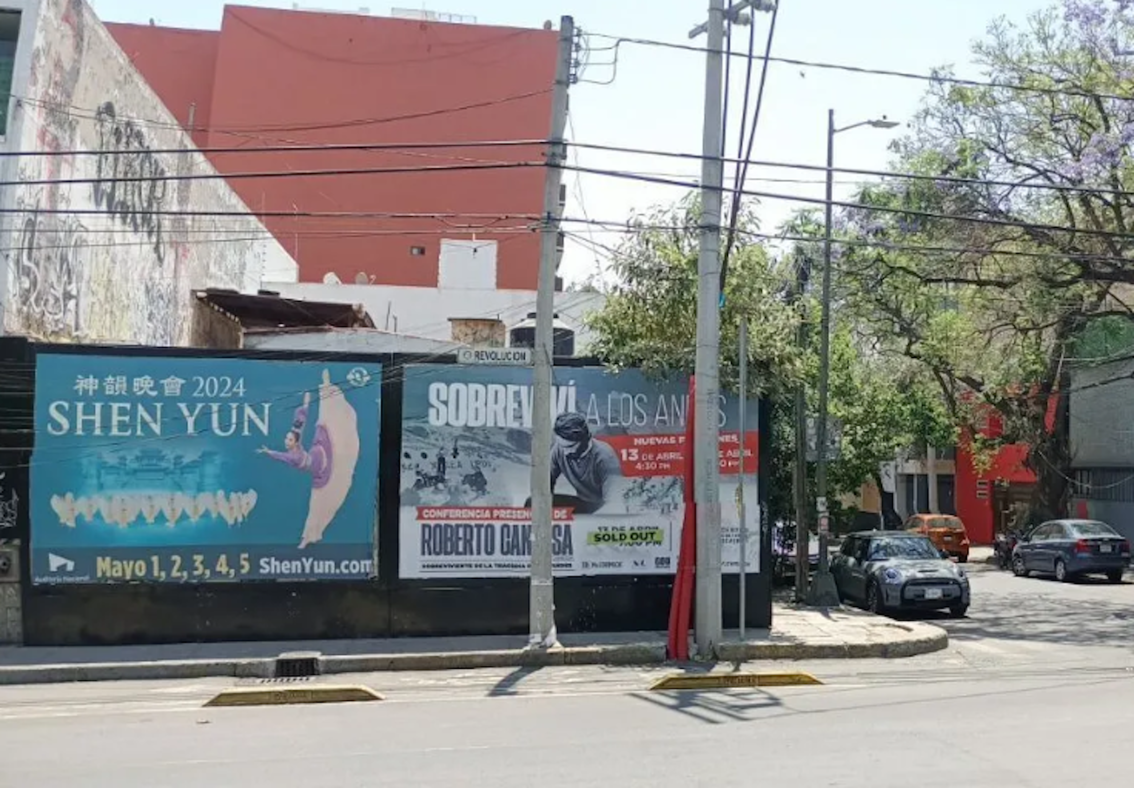 La imagen muestra una calle urbana con edificios y anuncios publicitarios. Se ven carteles promocionando eventos culturales como Shen Yun y una conferencia. Aunque no está directamente relacionada con el sector inmobiliario, la escena urbana refleja el uso del espacio público para publicidad y la mezcla de estructuras comerciales y residenciales típica de áreas urbanas.