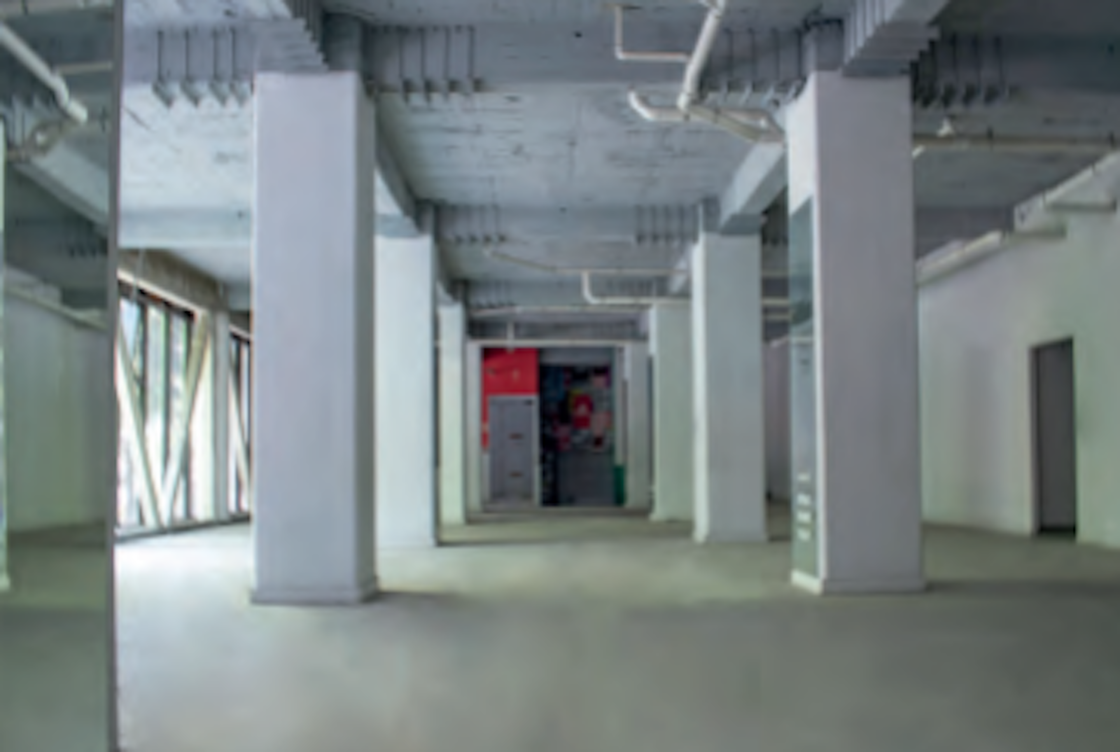 La imagen muestra el interior de un edificio en construcción o renovación. Se aprecia un amplio espacio con columnas blancas, techos altos con instalaciones expuestas y paredes sin acabar. Al fondo se ve una puerta roja y algunos elementos coloridos, posiblemente señalización. El espacio parece estar preparado para ser dividido en oficinas o locales comerciales, típico de desarrollos inmobiliarios en fase de obra gris.