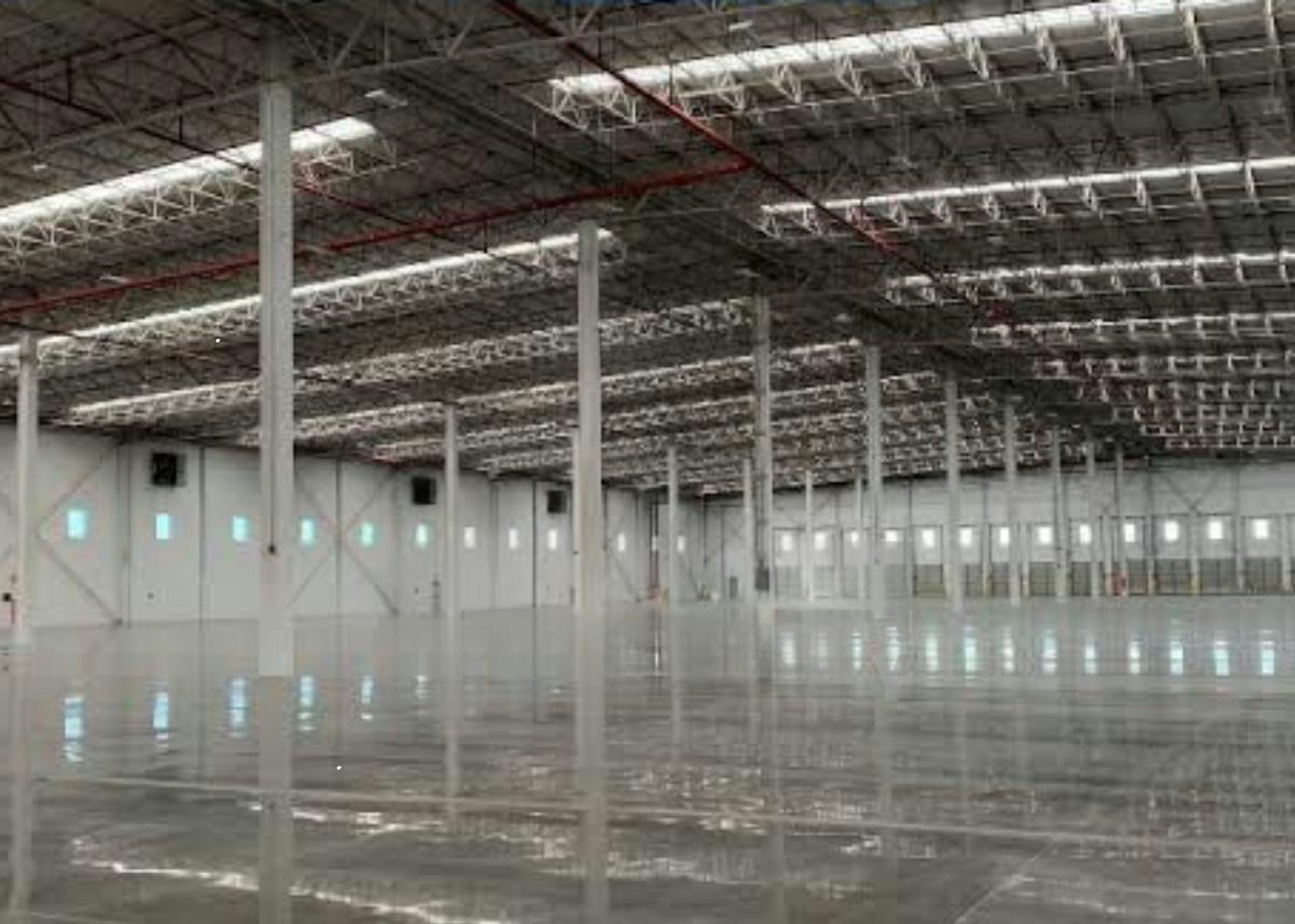 La imagen muestra un amplio almacén o centro de distribución industrial con un techo alto y estructuras metálicas. El suelo reflectante sugiere un acabado pulido o recién instalado. Se ve el logotipo y texto de Strategic Industry Logistics superpuesto, indicando que es una instalación logística. Las luces brillantes y el espacio abierto sugieren un diseño moderno y eficiente para operaciones de almacenamiento y distribución a gran escala.