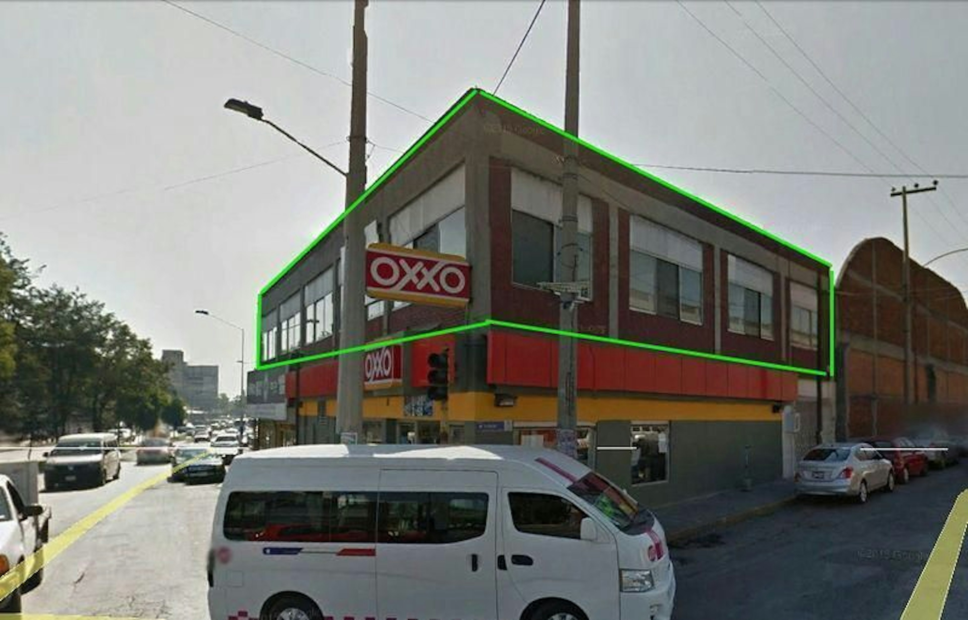 La imagen muestra un edificio de esquina de dos pisos que alberga una tienda OXXO, una cadena de tiendas de conveniencia muy común en México. El edificio está destacado con un contorno verde y tiene una fachada de ladrillo rojo con grandes ventanales. La tienda ocupa la planta baja con sus característicos colores rojo y amarillo. La ubicación parece ser una zona urbana con tráfico y otros negocios alrededor.