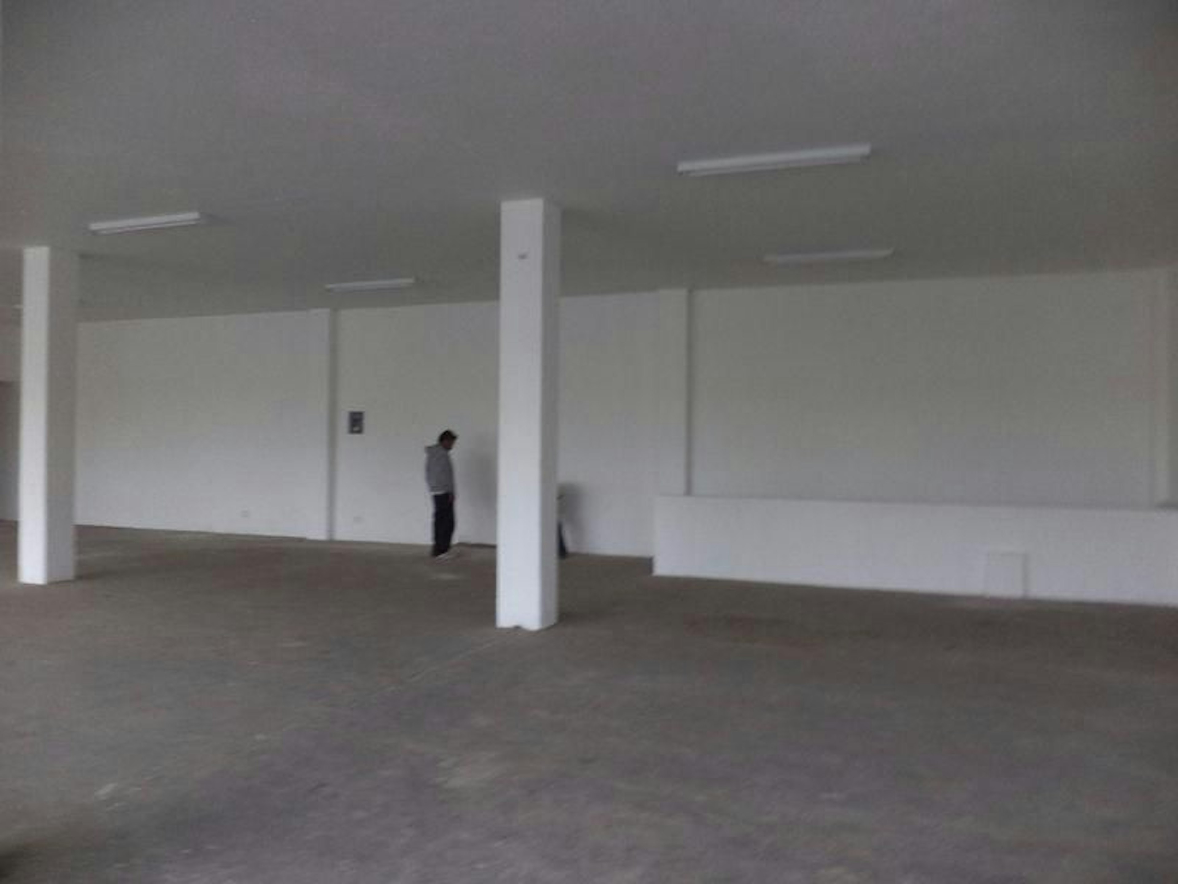 La imagen muestra un espacio interior amplio y vacío, posiblemente un local comercial o área de oficinas en construcción o renovación. Las paredes son blancas y lisas, con columnas visibles. El techo tiene luces fluorescentes. El suelo parece ser de concreto sin acabar. Hay una figura solitaria en el fondo, lo que da una idea de la escala del espacio. El ambiente general es austero y minimalista, típico de propiedades comerciales en fase de desarrollo.