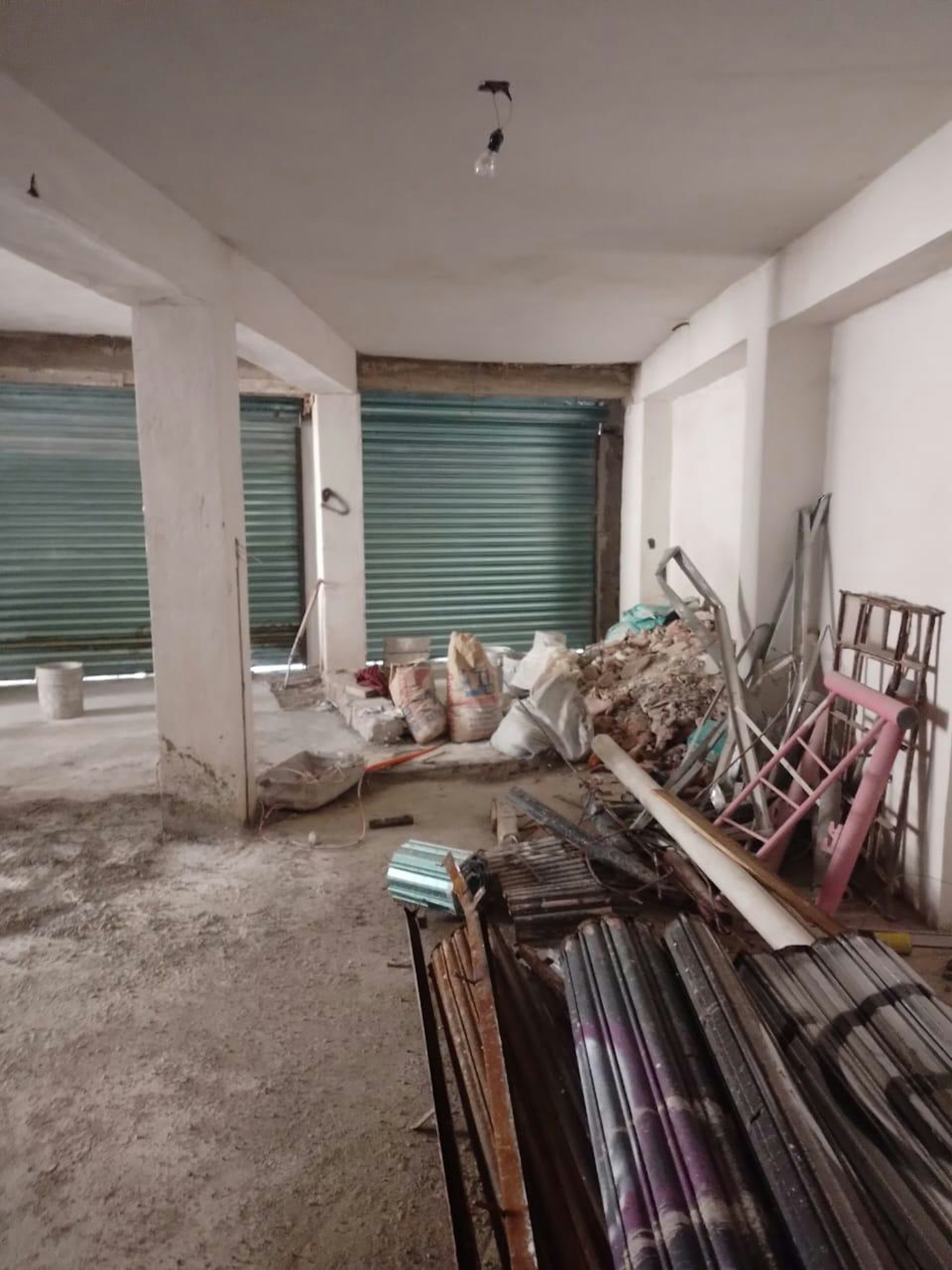 La imagen muestra un espacio interior en proceso de renovación o construcción. Hay escombros, bolsas de materiales y herramientas dispersas por el suelo. Se ven columnas blancas, un techo sin terminar y puertas enrollables de metal verde. El espacio parece ser un garaje o área de almacenamiento en renovación, con diversos materiales de construcción apilados, incluyendo tuberías y estructuras metálicas.