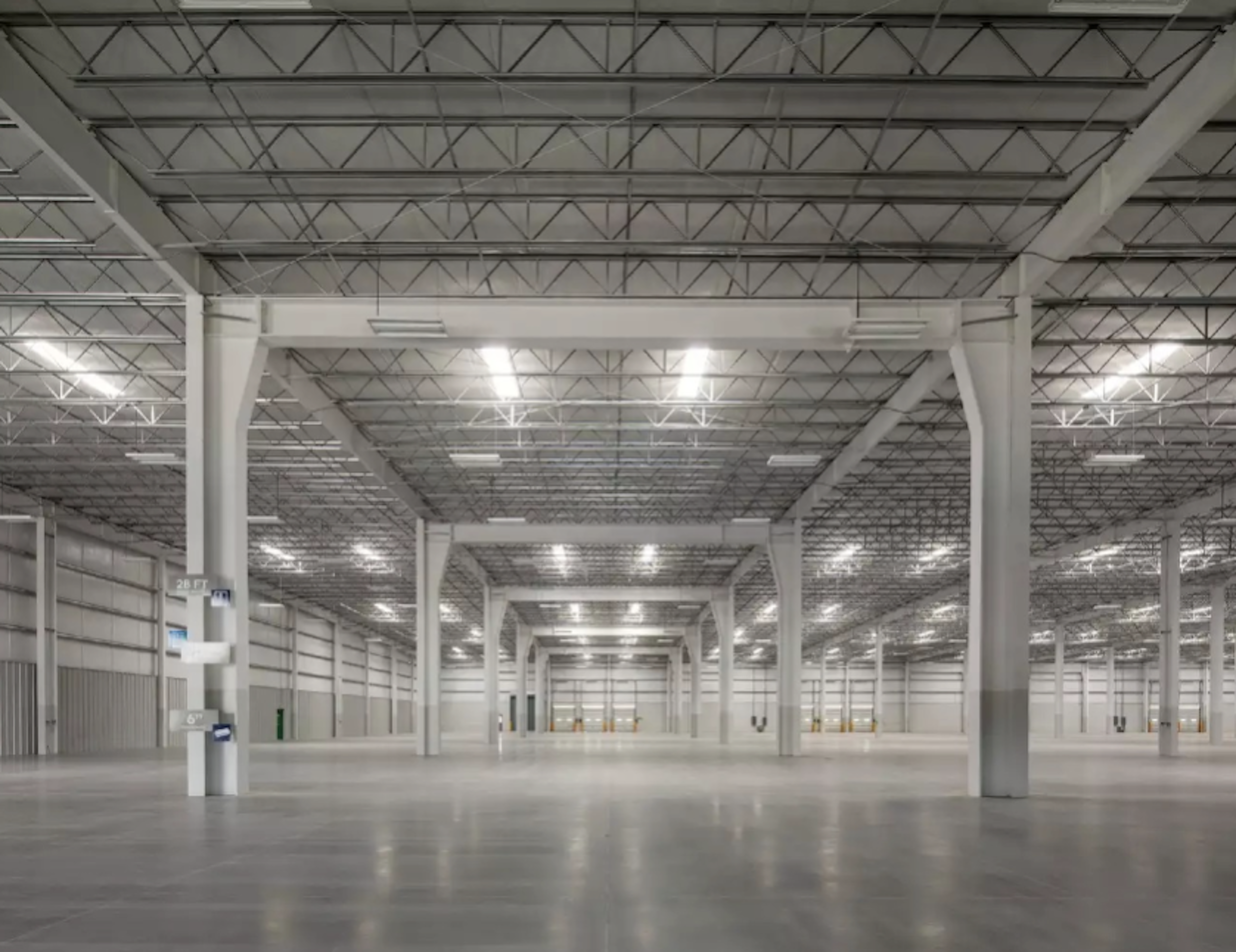 La imagen muestra un vasto espacio interior industrial, típico de un almacén o centro de distribución moderno. El techo alto está sostenido por una estructura metálica visible. Las paredes y el suelo son de color claro, creando un ambiente luminoso y espacioso. La iluminación artificial está distribuida uniformemente, resaltando la amplitud del lugar. Este tipo de espacio es ideal para operaciones logísticas o almacenamiento a gran escala en el sector inmobiliario industrial.