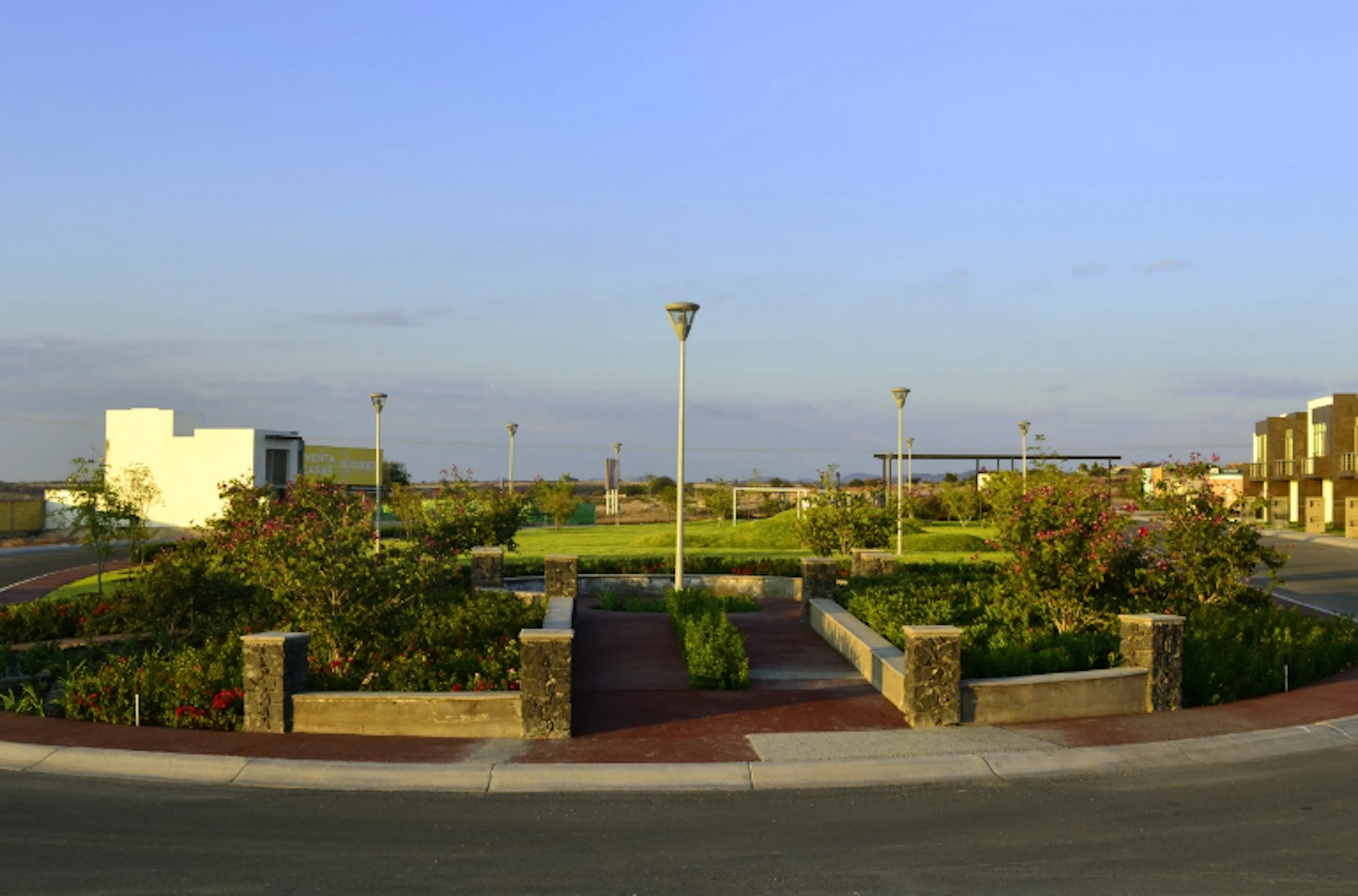 La imagen muestra un desarrollo inmobiliario moderno con áreas verdes bien cuidadas. Se aprecia un jardín central con caminos, plantas y arbustos floridos. Alrededor hay edificios residenciales de baja altura y farolas. El cielo es azul claro, sugiriendo un clima agradable. El diseño paisajístico y la arquitectura indican un complejo residencial de calidad con énfasis en espacios comunes atractivos.