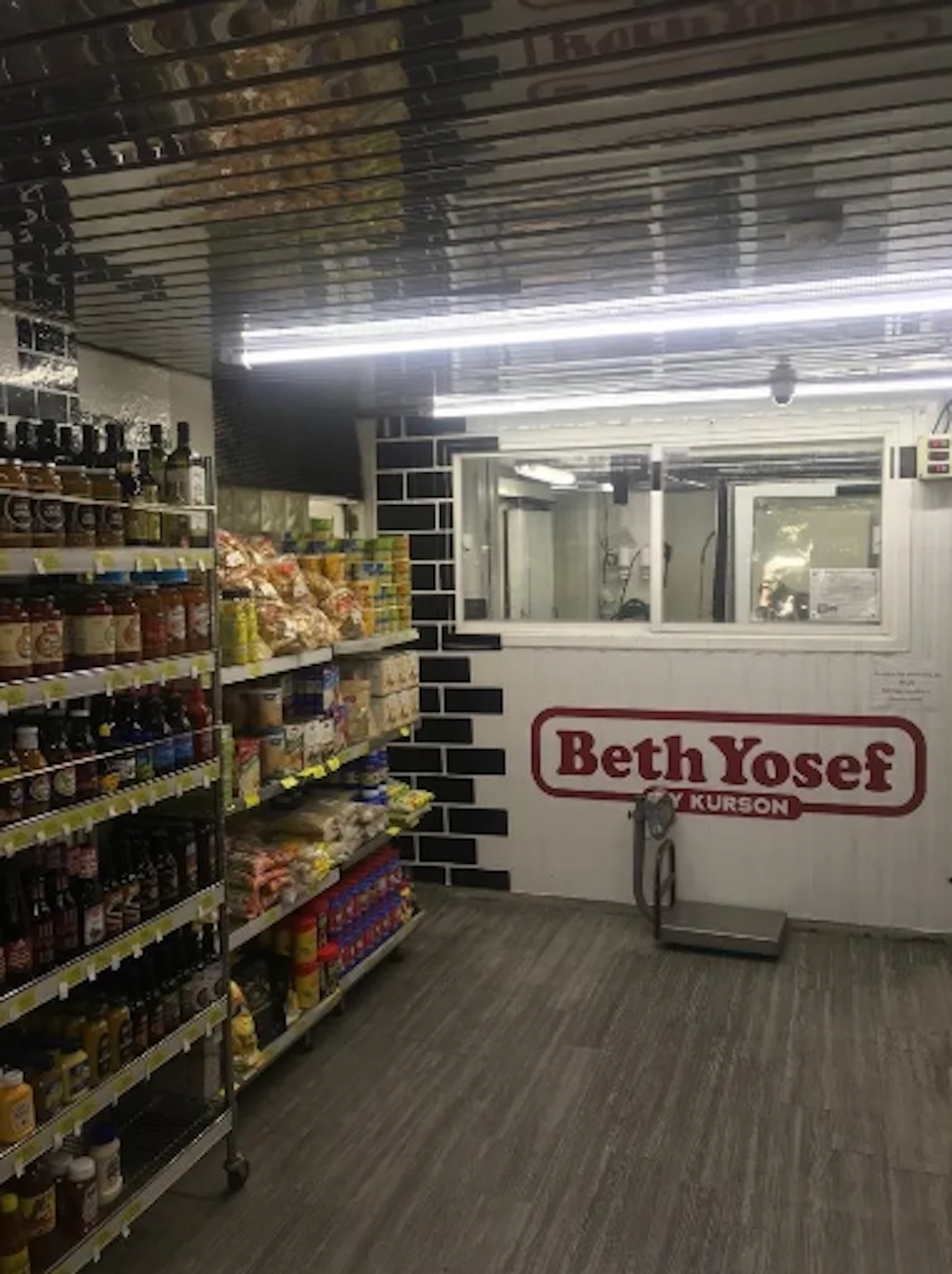 La imagen muestra el interior de una tienda de comestibles llamada Beth Yosef. Se observan estanterías llenas de productos alimenticios, botellas y otros artículos. Al fondo hay una ventana de servicio con el logo de la tienda. El espacio tiene iluminación fluorescente y un suelo de madera, dando una apariencia moderna y limpia a pesar de ser un local pequeño.