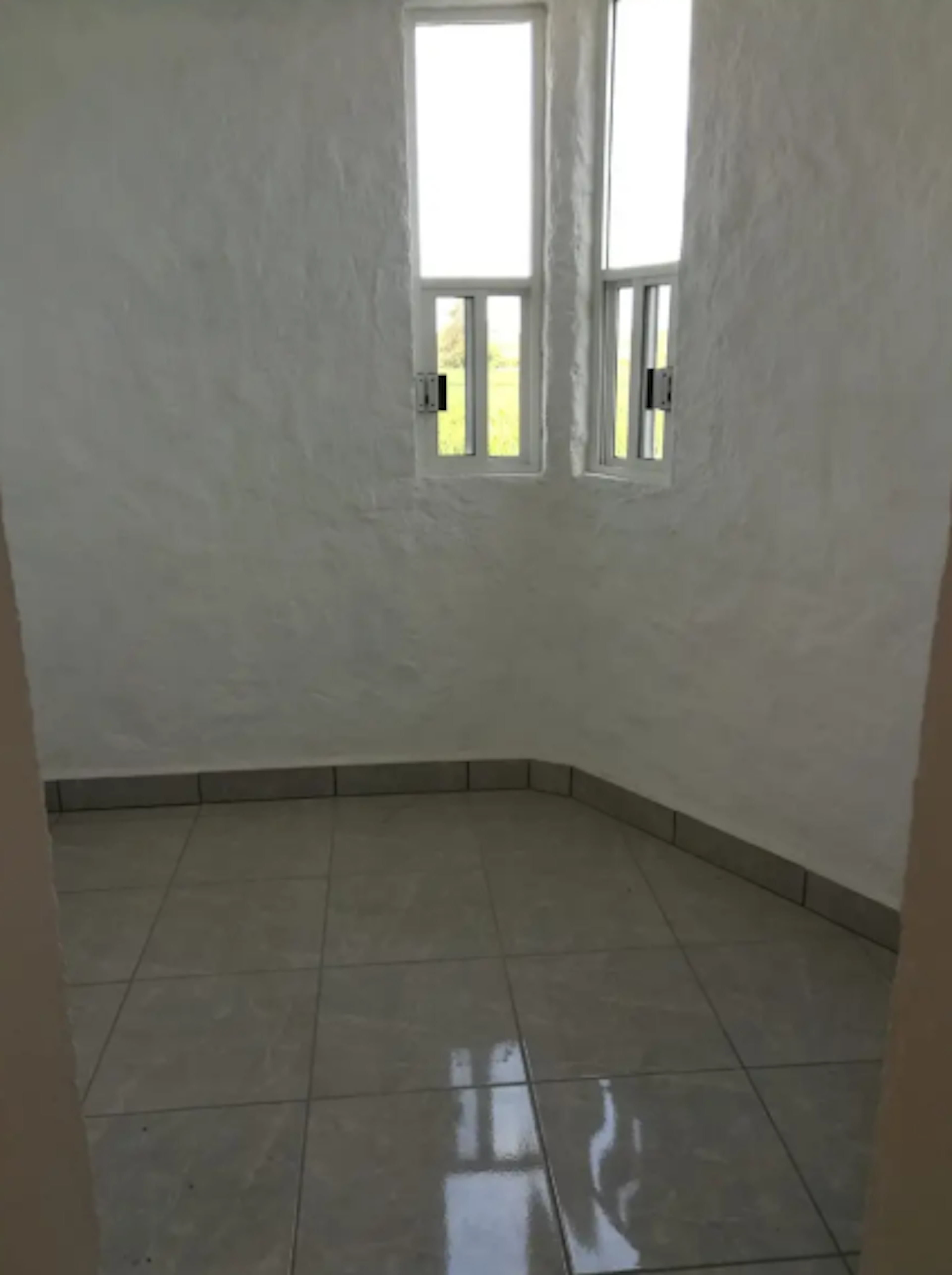 La imagen muestra un espacio interior vacío con paredes blancas y piso de baldosas. Hay una ventana estrecha y alta que permite la entrada de luz natural. El espacio parece ser una habitación pequeña o un rincón de una casa, posiblemente en construcción o recién renovada. Las paredes tienen una textura rugosa y el piso refleja la luz, indicando que está limpio o recién instalado.