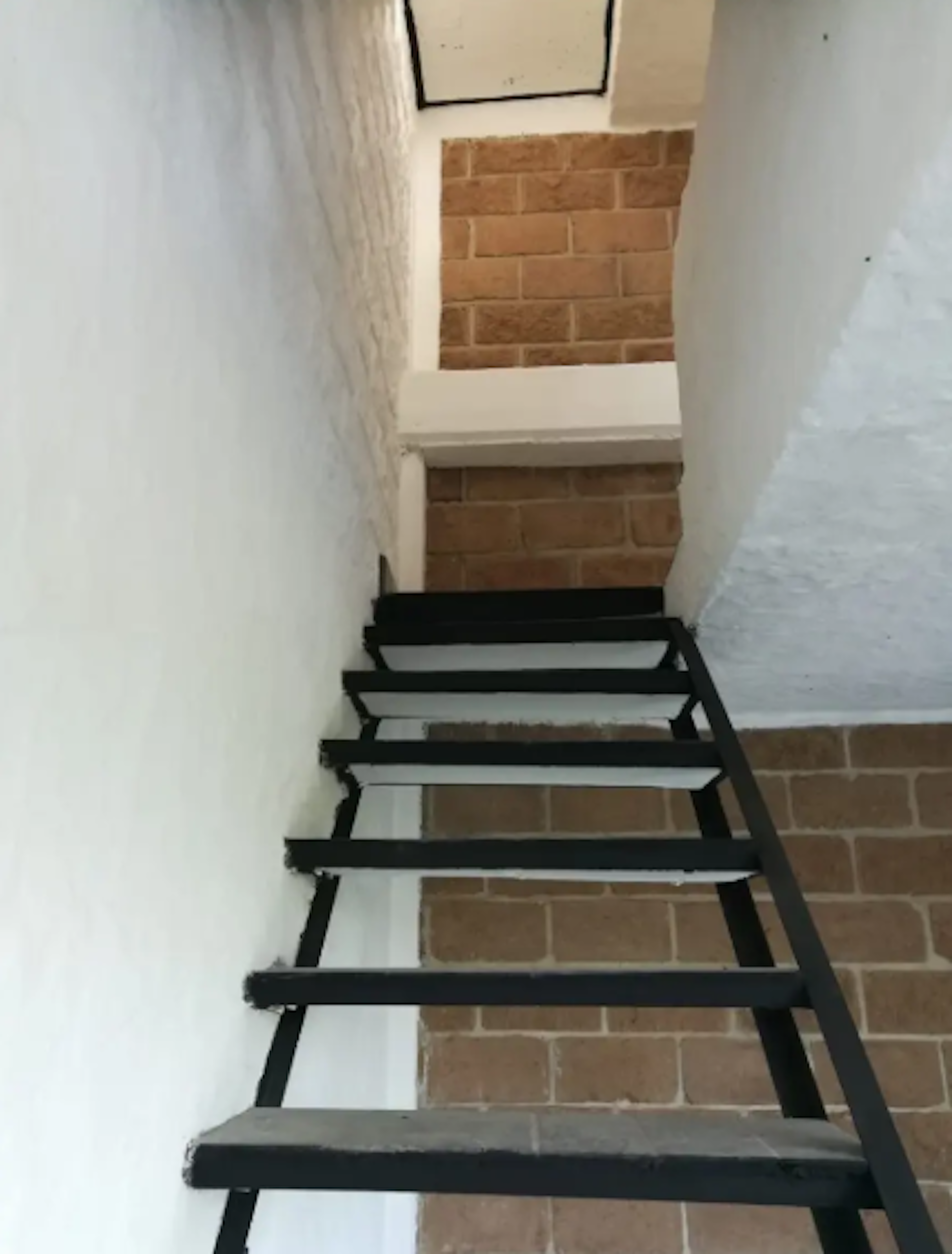 La imagen muestra una escalera interior en construcción. Es una estructura metálica negra sin barandillas, con peldaños provisionales blancos. Las paredes alrededor son blancas con secciones de ladrillo expuesto. El espacio es estrecho y parece ser parte de una renovación o construcción en progreso de una vivienda o edificio residencial.