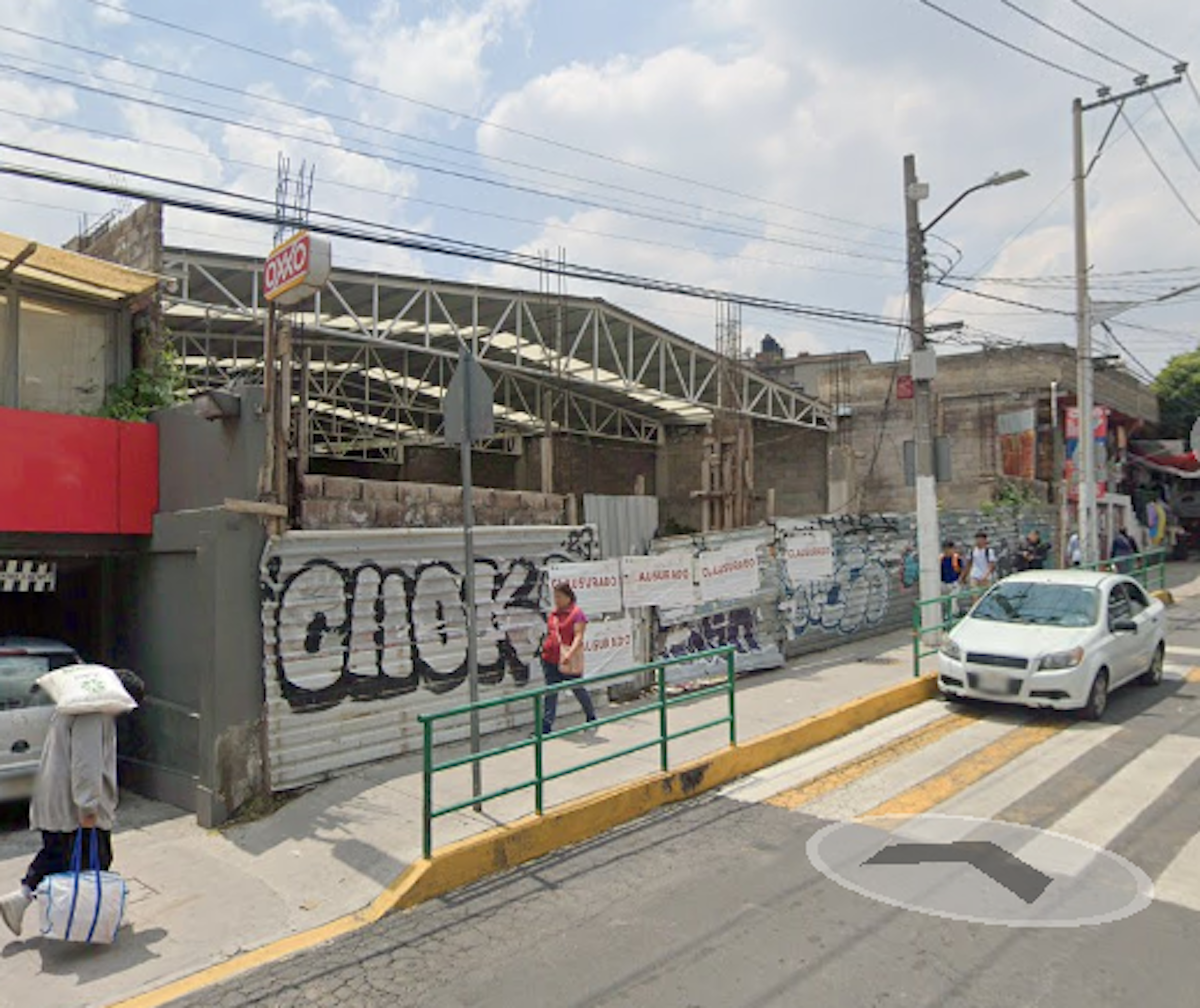 La imagen muestra una calle urbana con una construcción en progreso. Se ve una estructura metálica de un edificio en construcción, junto a un muro con grafitis. Hay una tienda Oxxo visible, cables eléctricos aéreos y postes. La calle tiene un cruce peatonal y barandillas. Se observan algunos peatones y un automóvil. El entorno sugiere una zona en desarrollo o remodelación urbana.