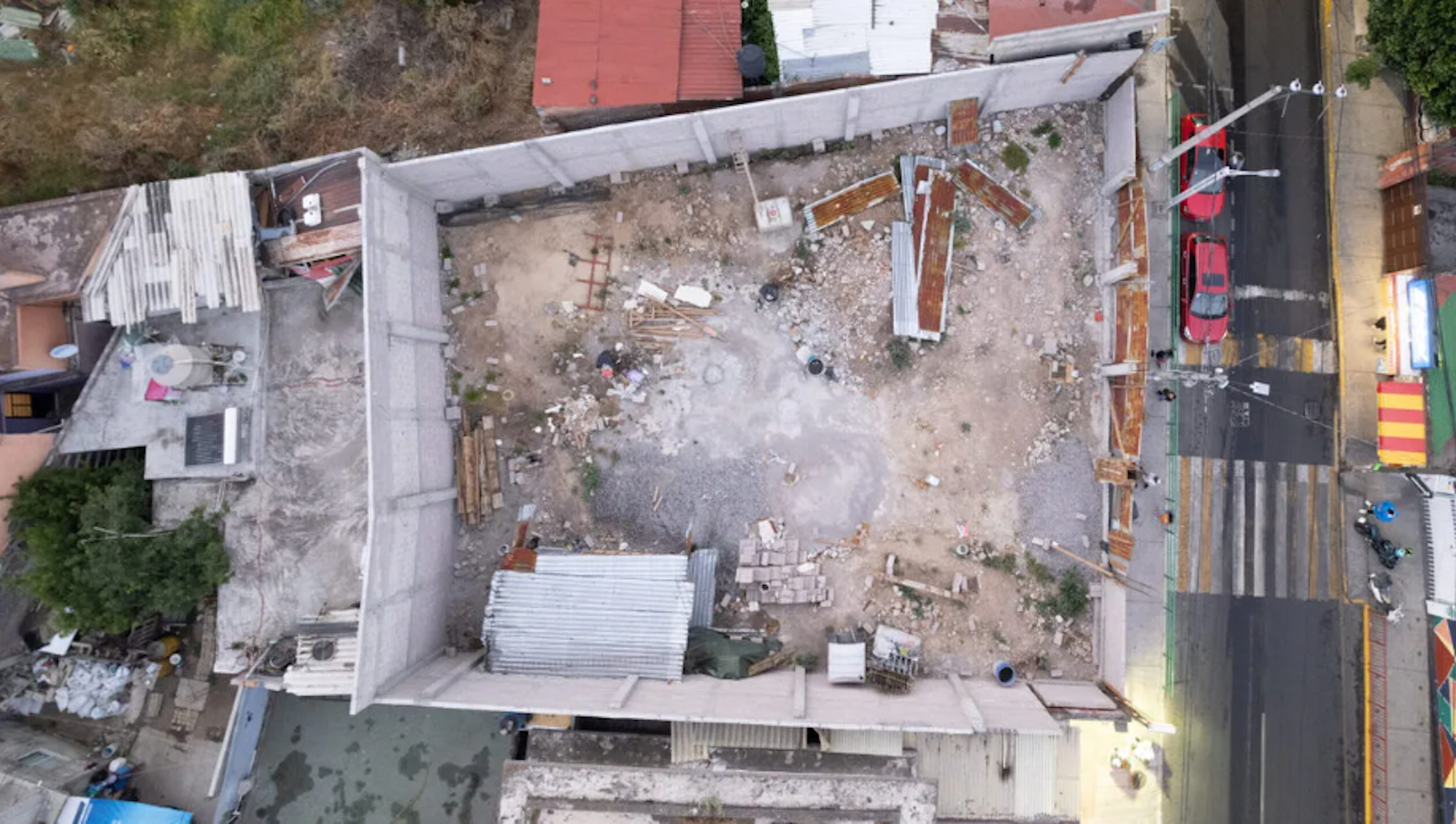 La imagen muestra una vista aérea de un sitio de construcción o demolición en una zona urbana. Se observa un área rectangular con escombros, materiales de construcción dispersos y algunas estructuras parciales. Alrededor hay edificios y calles, lo que sugiere que es un proyecto de desarrollo o renovación en curso en medio de un área poblada. El estado del sitio indica que está en una fase temprana de construcción o en proceso de limpieza.