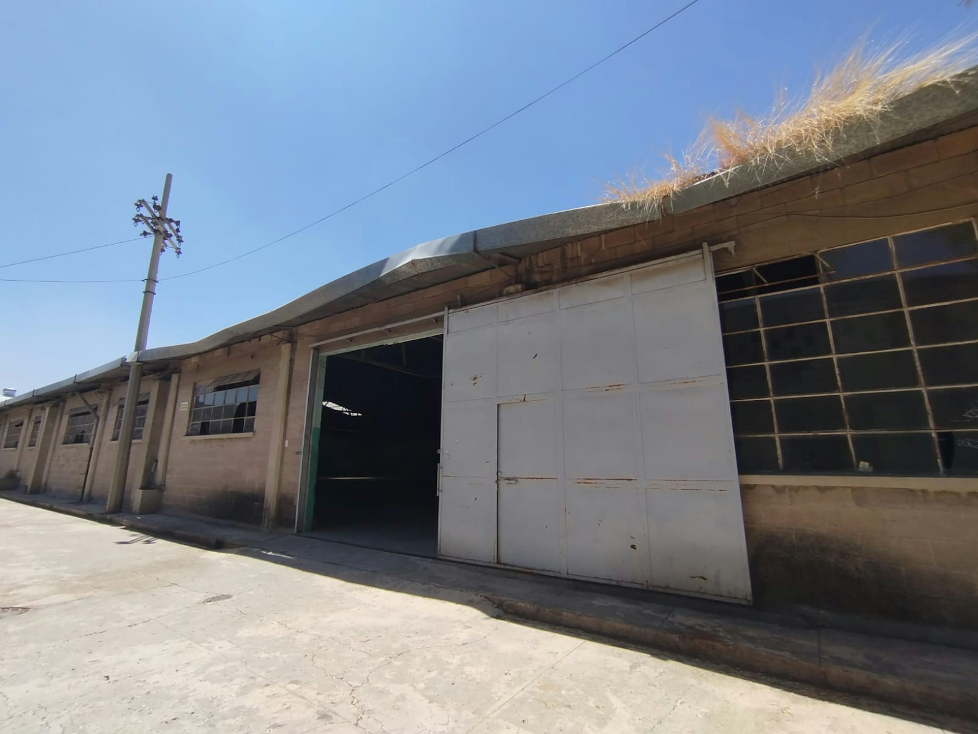 La imagen muestra un edificio industrial o almacén en estado deteriorado. Tiene paredes de concreto, una gran puerta blanca metálica y ventanas en mal estado. El techo parece estar cubierto de paja o vegetación. Se observa un poste eléctrico cercano. El entorno sugiere una zona industrial o rural. La estructura parece abandonada o en desuso, lo que podría representar una oportunidad para renovación o redevelopment en el sector inmobiliario.
