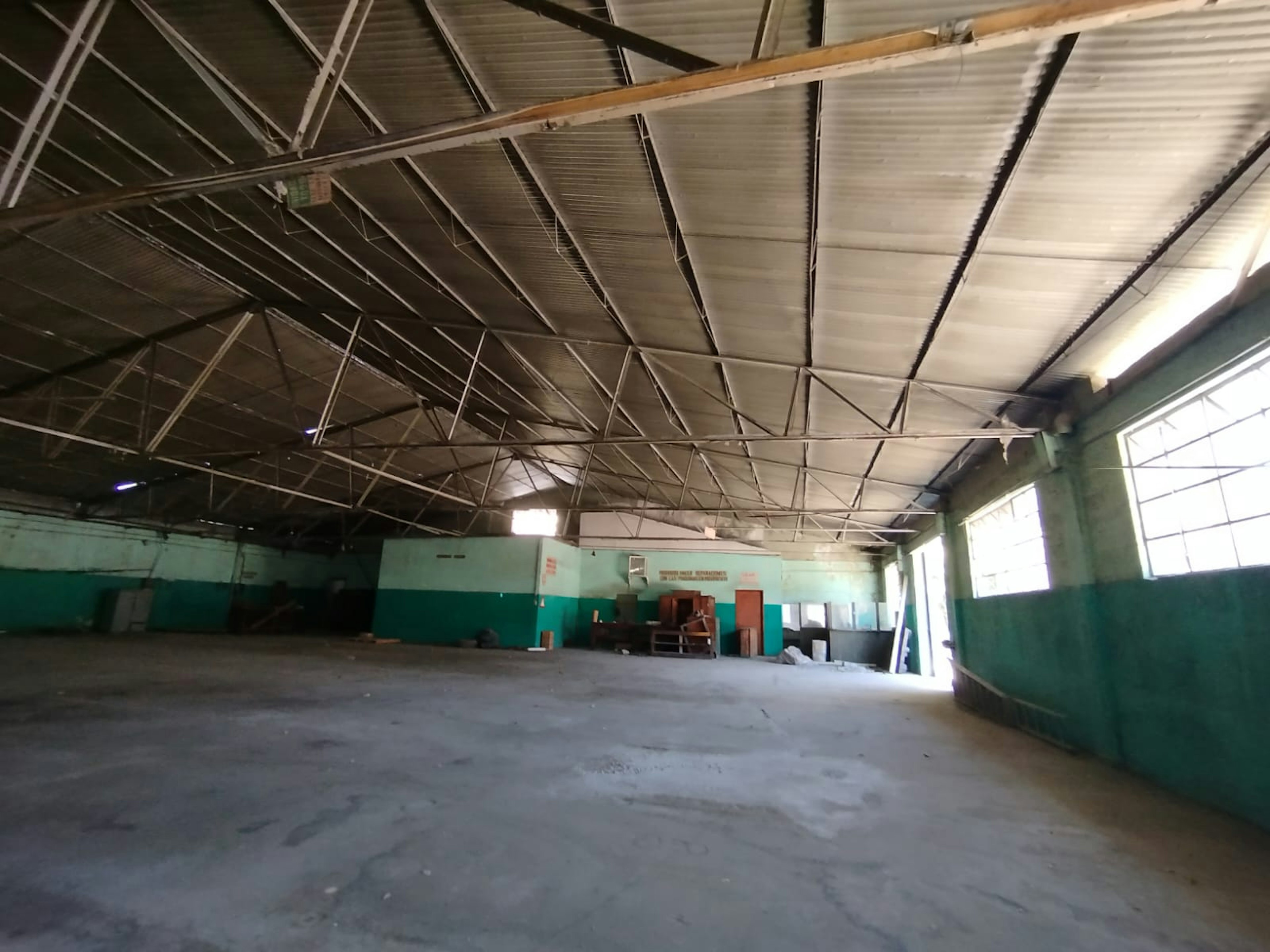 Esta imagen muestra un amplio espacio interior industrial o comercial abandonado. Tiene un techo alto con estructura metálica visible, paredes pintadas en verde y turquesa, y un piso de concreto. El lugar parece estar en desuso, con algunos muebles o escombros dispersos. Las ventanas altas permiten la entrada de luz natural. El espacio podría ser adecuado para rehabilitación o reconversión en un loft, almacén o espacio comercial.