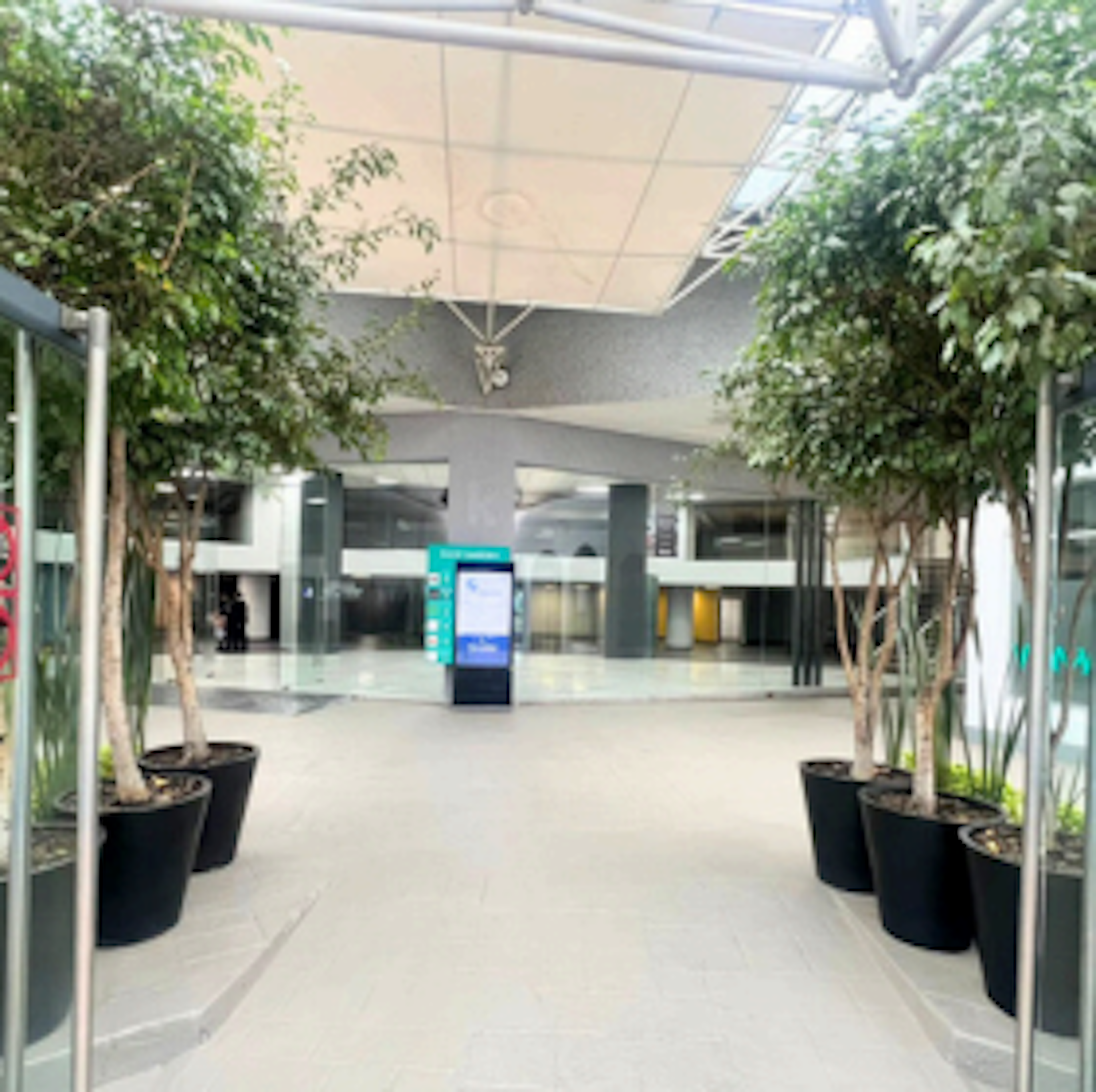 La imagen muestra un moderno y espacioso vestíbulo de un edificio corporativo o centro comercial. Cuenta con un techo alto con paneles, plantas decorativas en macetas negras a lo largo de los lados, y un kiosco digital interactivo en el centro. El espacio tiene un diseño limpio y minimalista con tonos blancos y grises, creando una atmósfera acogedora y profesional.