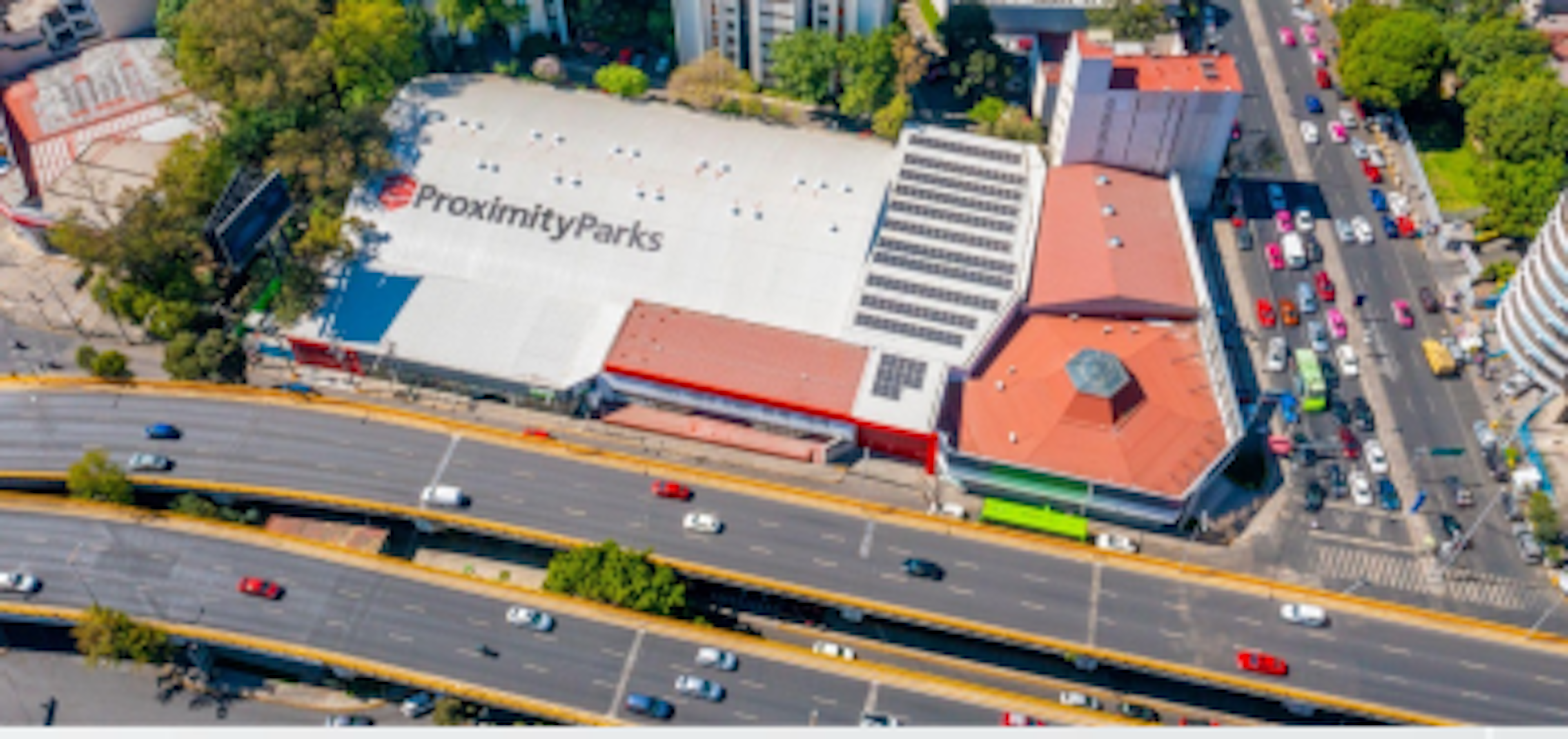 La imagen muestra una vista aérea de un área urbana con un gran edificio etiquetado como ProximityParks. Este parece ser un centro comercial o complejo de tiendas con un amplio techo blanco y paneles solares. Está rodeado de calles concurridas con múltiples carriles de tráfico, otros edificios comerciales y áreas verdes. La ubicación parece ser una zona comercial activa en una ciudad grande.