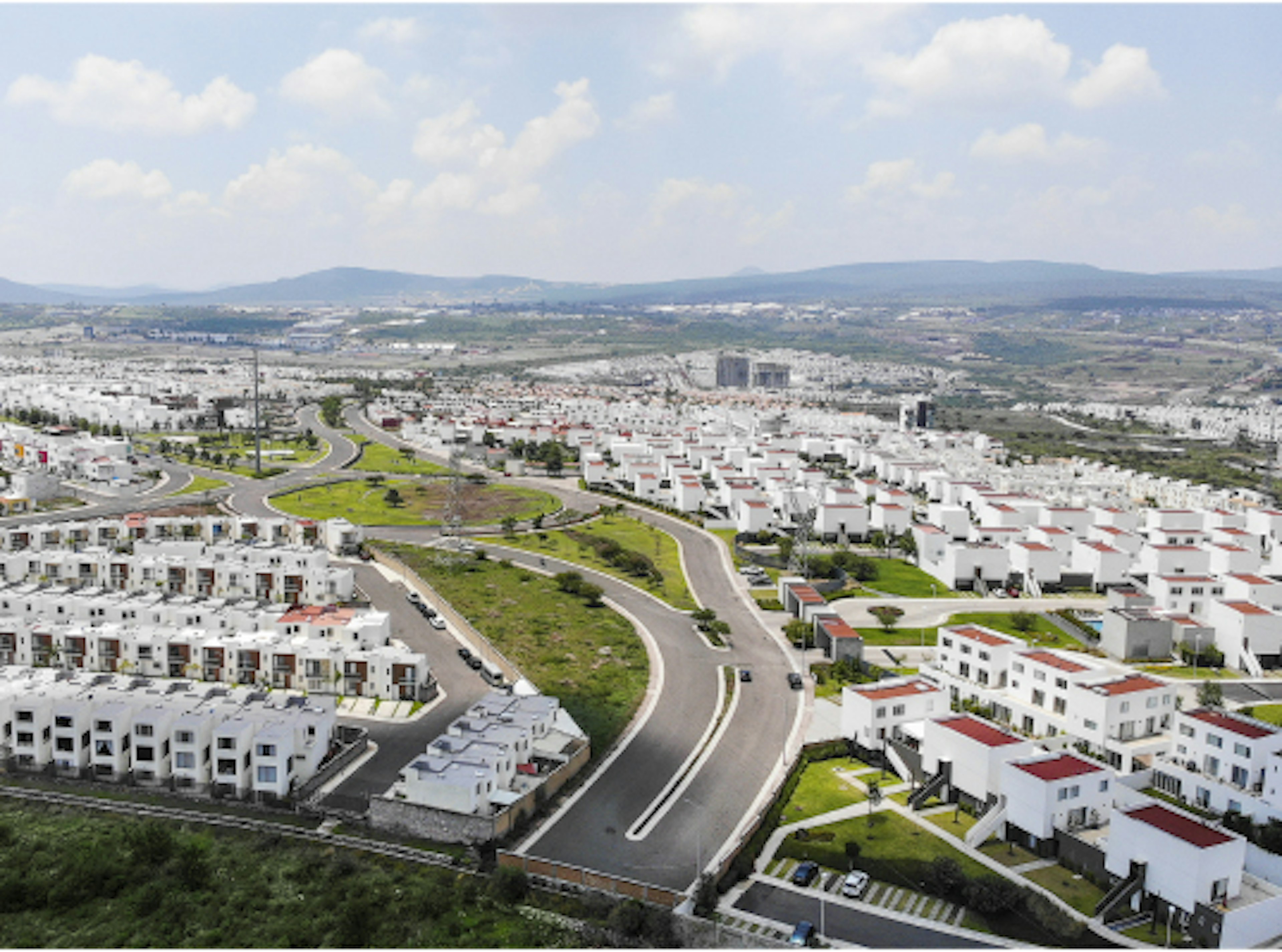 La imagen muestra una vista aérea de un extenso desarrollo inmobiliario con múltiples casas y edificios de color blanco con techos rojos. Se observa una red de calles curvas que serpentean entre las viviendas, creando un patrón ordenado. El paisaje urbano se extiende hasta el horizonte, donde se ven montañas distantes. La planificación urbana es evidente, con áreas verdes y espacios abiertos integrados en el diseño.