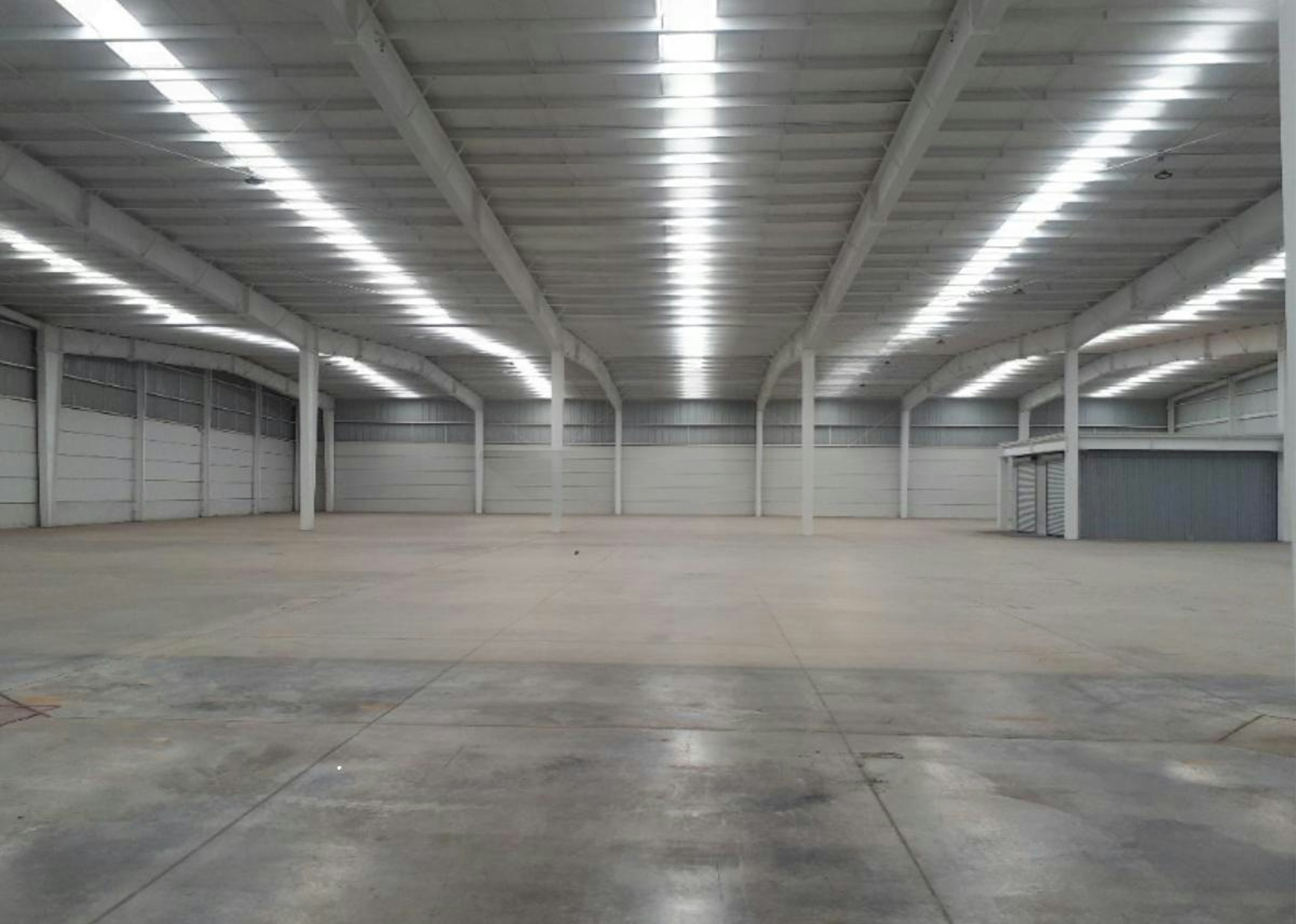 La imagen muestra un amplio espacio industrial vacío con techos altos y luminarias fluorescentes. Las paredes y el suelo son de concreto, con algunas marcas en el piso. Se observan columnas estructurales y puertas de acceso. El espacio parece ser una nave industrial o almacén logístico. En primer plano se aprecia un logo translúcido con el texto Strategic Industry Logistics Industry