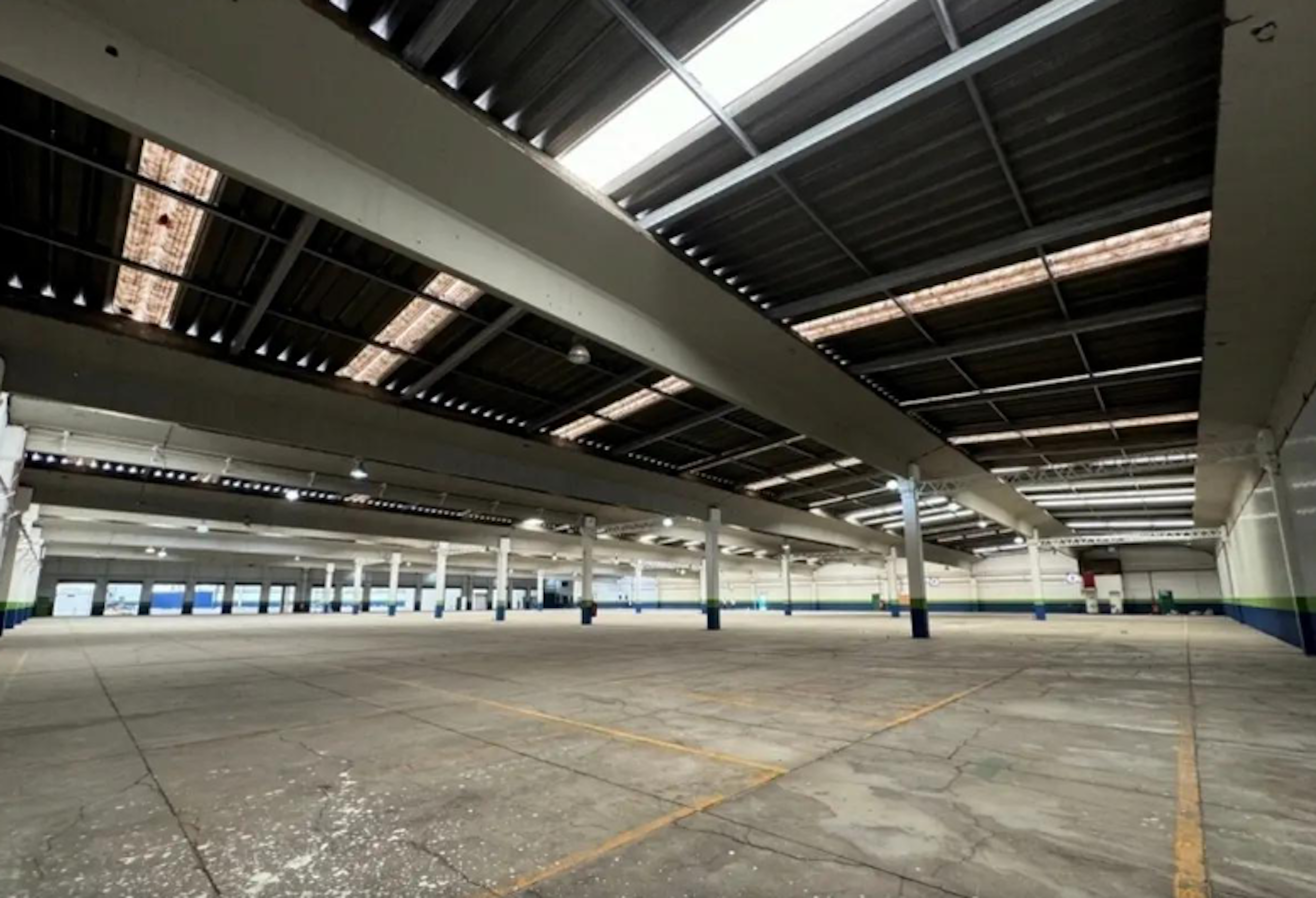 La imagen muestra un amplio espacio industrial o de almacenamiento vacío. Tiene un techo alto con vigas y tragaluces visibles. El suelo es de concreto con algunas marcas amarillas. Las columnas están pintadas de blanco con bases azules. El espacio parece estar bien iluminado con luces artificiales y luz natural. Es un área extensa que podría ser utilizada para almacenamiento, manufactura o ser convertida en un espacio comercial.