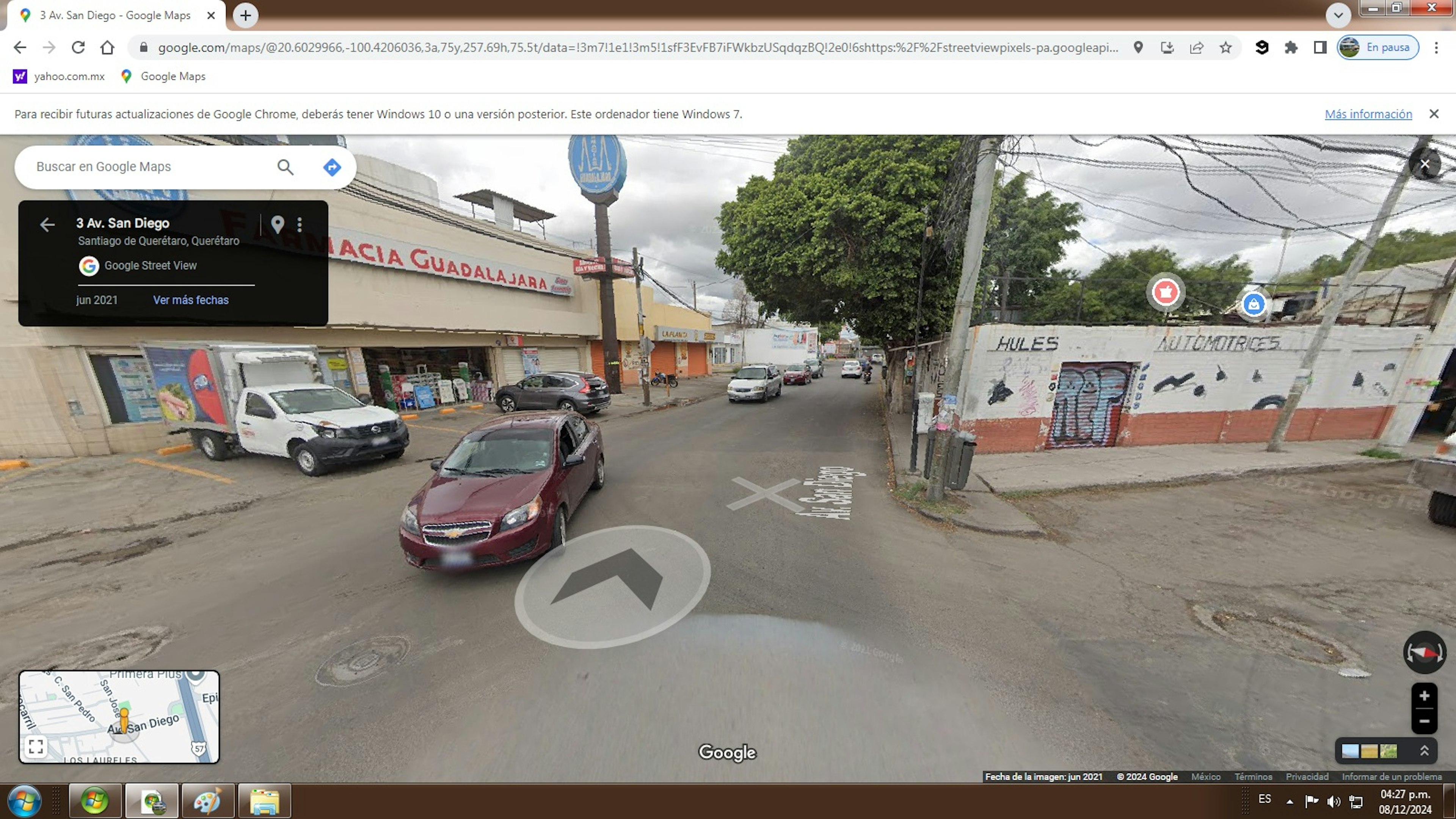 La imagen muestra una calle urbana en México con edificios comerciales y residenciales. Se ve una farmacia Guadalajara y un taller automotriz. La calle está pavimentada con algunos vehículos estacionados y en circulación. El área parece ser una zona de uso mixto con negocios y viviendas, típica de una ciudad mexicana de tamaño mediano. La infraestructura y el estado de los edificios sugieren una zona de clase media.