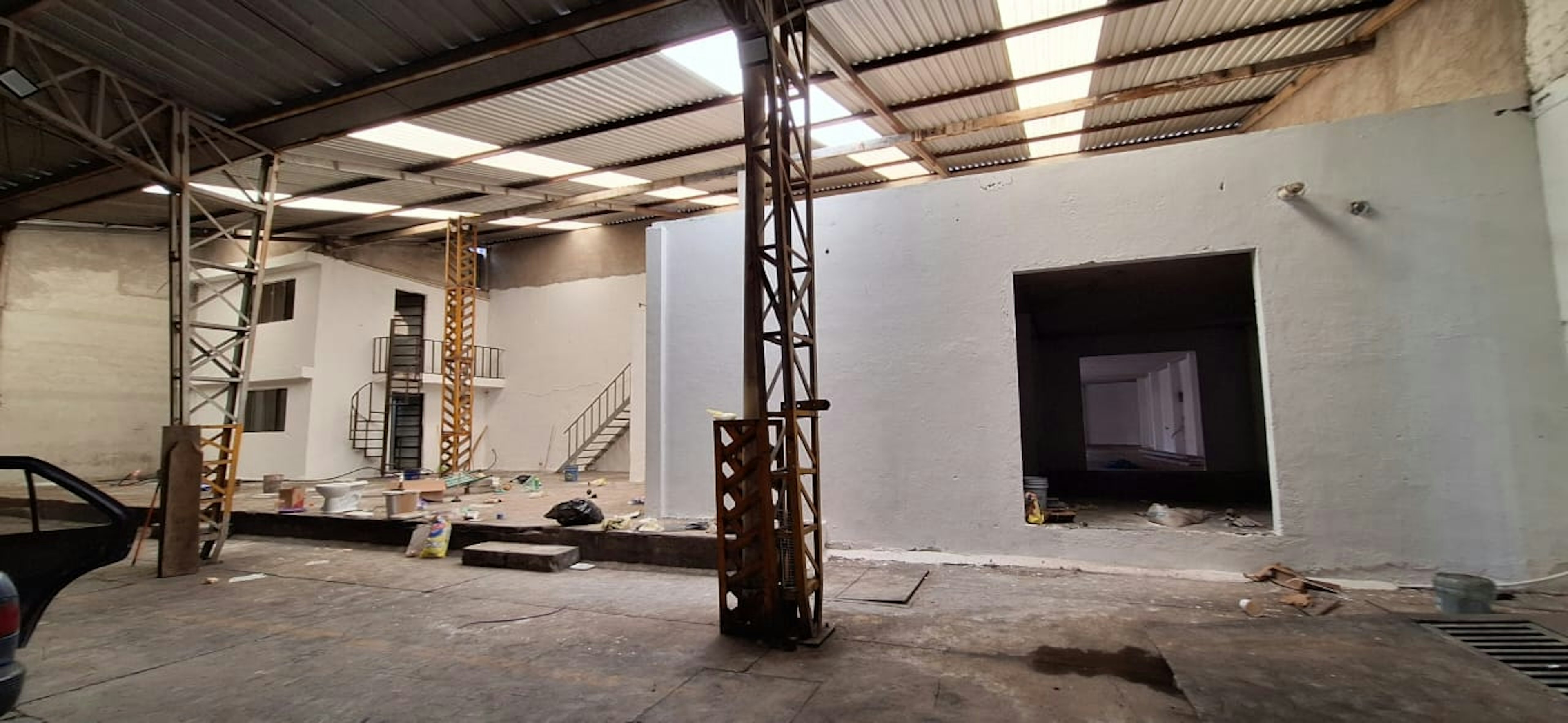 La imagen muestra un espacio industrial en proceso de renovación. Se observa una estructura con techo de lámina, columnas metálicas y paredes blancas recién pintadas. Hay escaleras visibles, una abertura grande que podría ser para una puerta de carga, y signos de trabajo en progreso como materiales de construcción dispersos. El espacio parece estar siendo convertido para un nuevo uso, posiblemente comercial o residencial.