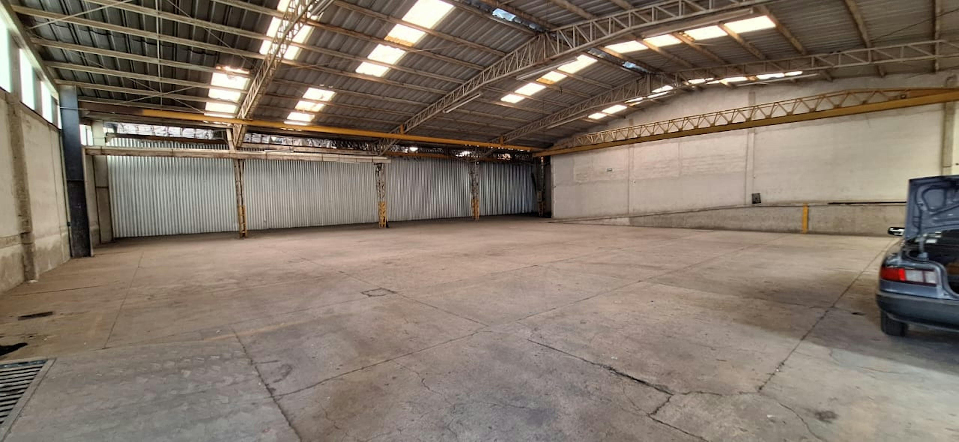 La imagen muestra un amplio espacio industrial vacío con techo alto y estructura metálica. El piso es de concreto y hay paredes de metal corrugado y concreto. Se observa iluminación natural a través de tragaluces y una parte de un vehículo en un extremo. El espacio parece ideal para almacenamiento, manufactura o como bodega, ofreciendo una gran área despejada para diversos usos comerciales o industriales.