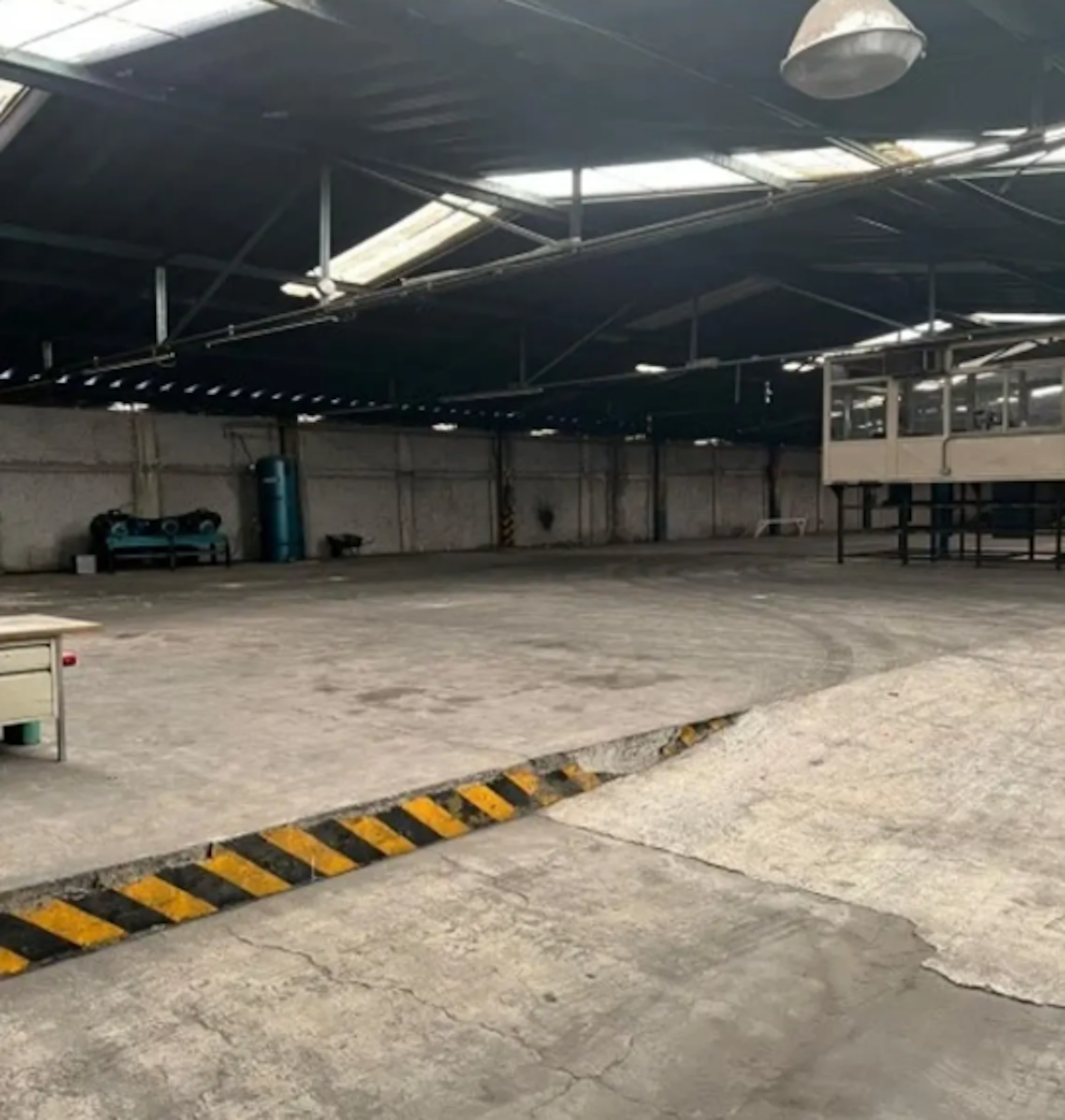 La imagen muestra un amplio espacio industrial vacío, posiblemente un almacén o nave industrial. Tiene un techo alto con estructura metálica y lucernarios. El suelo es de concreto con algunas grietas y una franja de seguridad amarilla y negra. Se observan paredes de concreto, algunas instalaciones como tuberías y un panel eléctrico. El espacio parece estar en desuso o listo para ser ocupado, ideal para almacenamiento o manufactura.