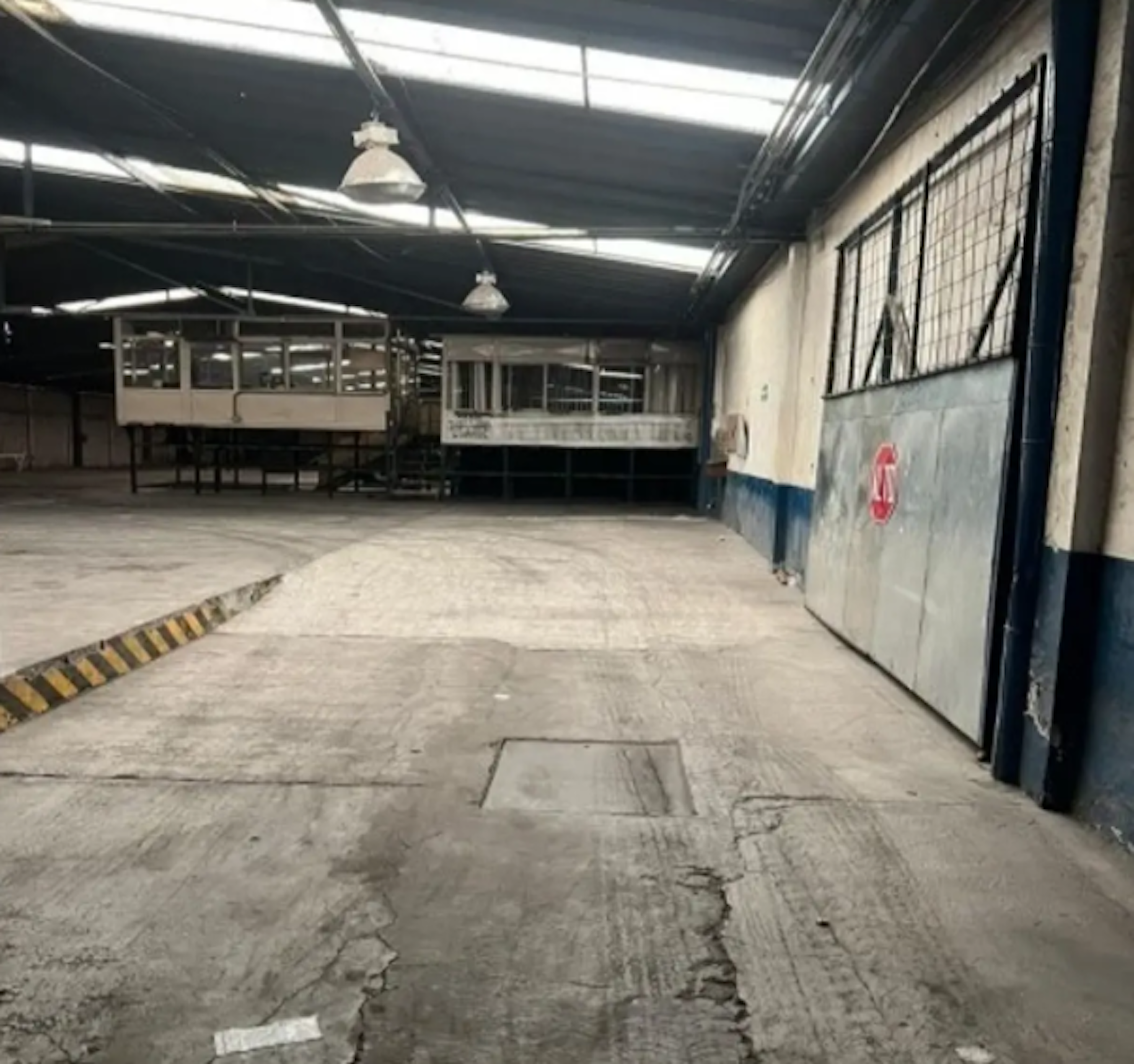 La imagen muestra un espacio industrial amplio y vacío, con techo alto y luces colgantes. Hay una estructura elevada con ventanas al fondo, posiblemente una oficina. El suelo es de concreto, con algunas grietas y marcas. Las paredes están parcialmente pintadas en azul y blanco, con una marca roja visible. El lugar parece ser un almacén o taller en desuso, potencialmente reutilizable para diversos fines comerciales o industriales.