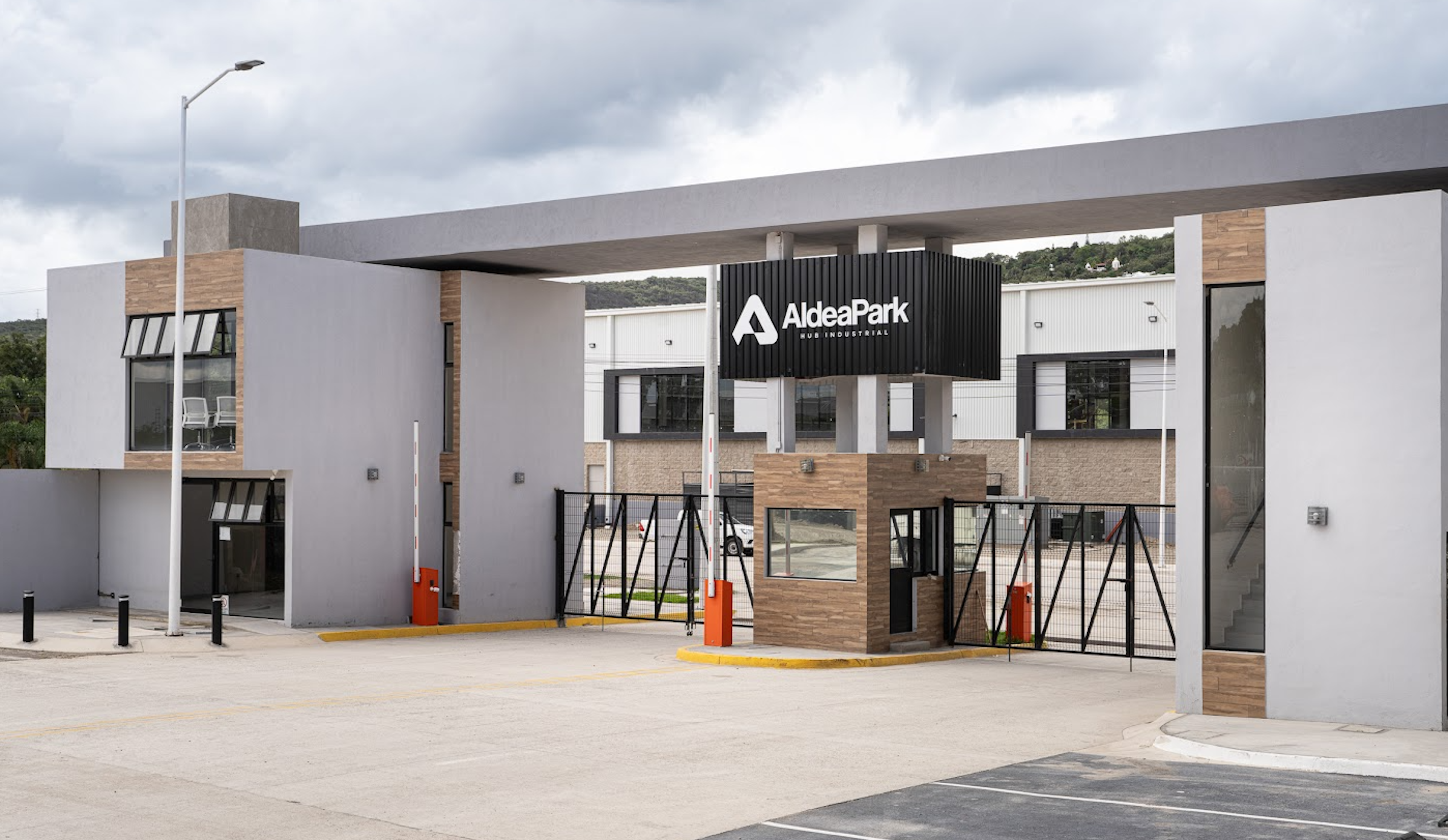 La imagen muestra la entrada moderna de un complejo llamado AldeaPark. La fachada combina elementos de concreto, vidrio y madera, con un diseño contemporáneo. Se aprecia una caseta de seguridad, barreras de acceso y un amplio espacio para vehículos. El letrero de AldeaPark destaca en negro sobre la estructura. El cielo nublado y la vegetación al fondo sugieren un entorno suburbano o periférico.