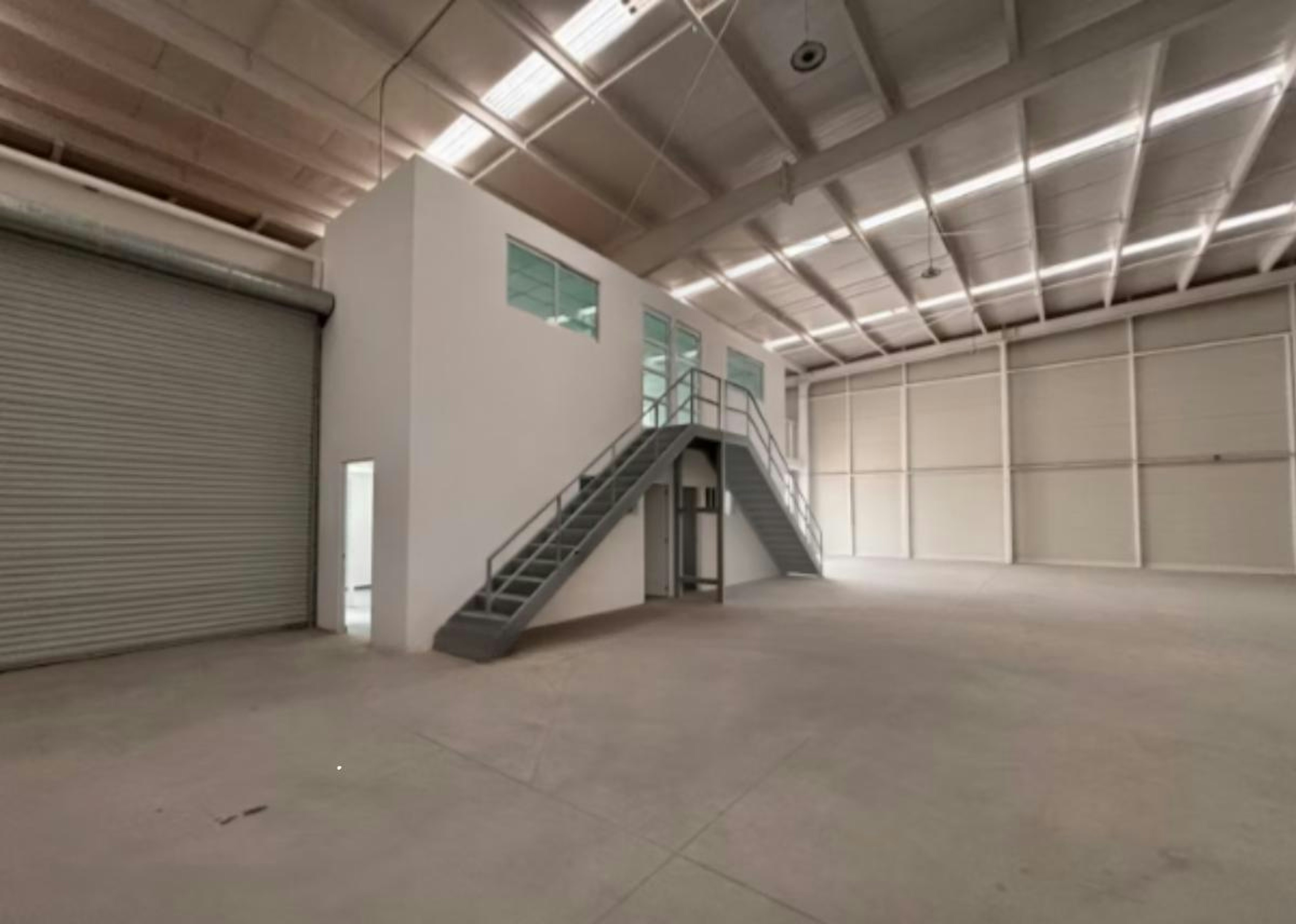 La imagen muestra un amplio espacio industrial interior con techos altos y luces fluorescentes. Hay una escalera metálica que conduce a un segundo nivel con ventanas. Se observa una puerta de garaje enrollable a la izquierda. El piso es de concreto y las paredes son blancas. La imagen tiene un logotipo superpuesto y texto que dice STRATEGIC INDUSTRY LOGISTIC INDUSTRY, sugiriendo que es una instalación logística o almacén.