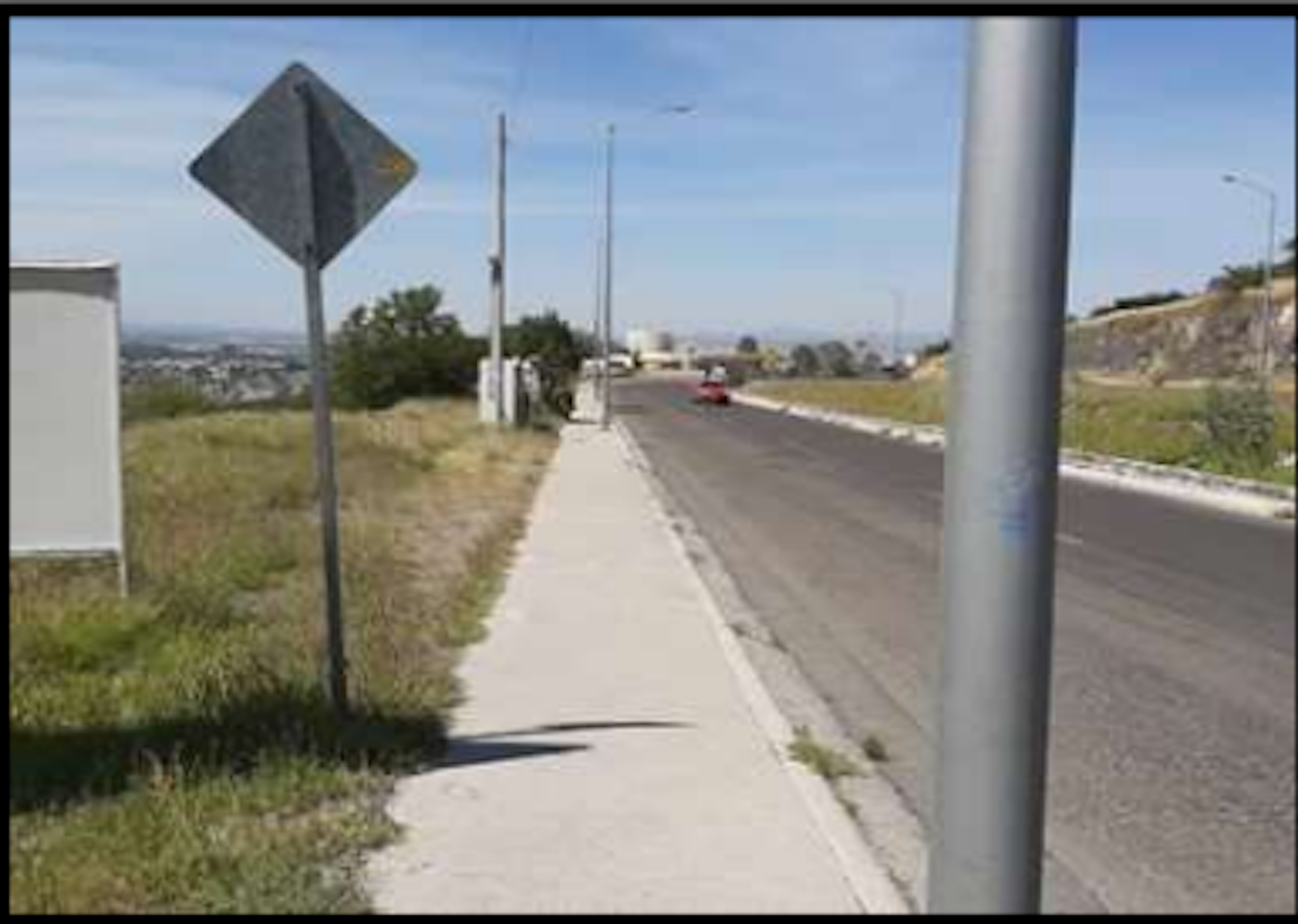 La imagen muestra una carretera suburbana con un paisaje urbano distante. Hay una acera estrecha junto a la calle, un letrero de tráfico y un poste de luz. El terreno circundante parece estar en desarrollo, con áreas de césped y vegetación dispersa. La vista sugiere una zona residencial en crecimiento o un área periférica de la ciudad con potencial para futuro desarrollo inmobiliario.