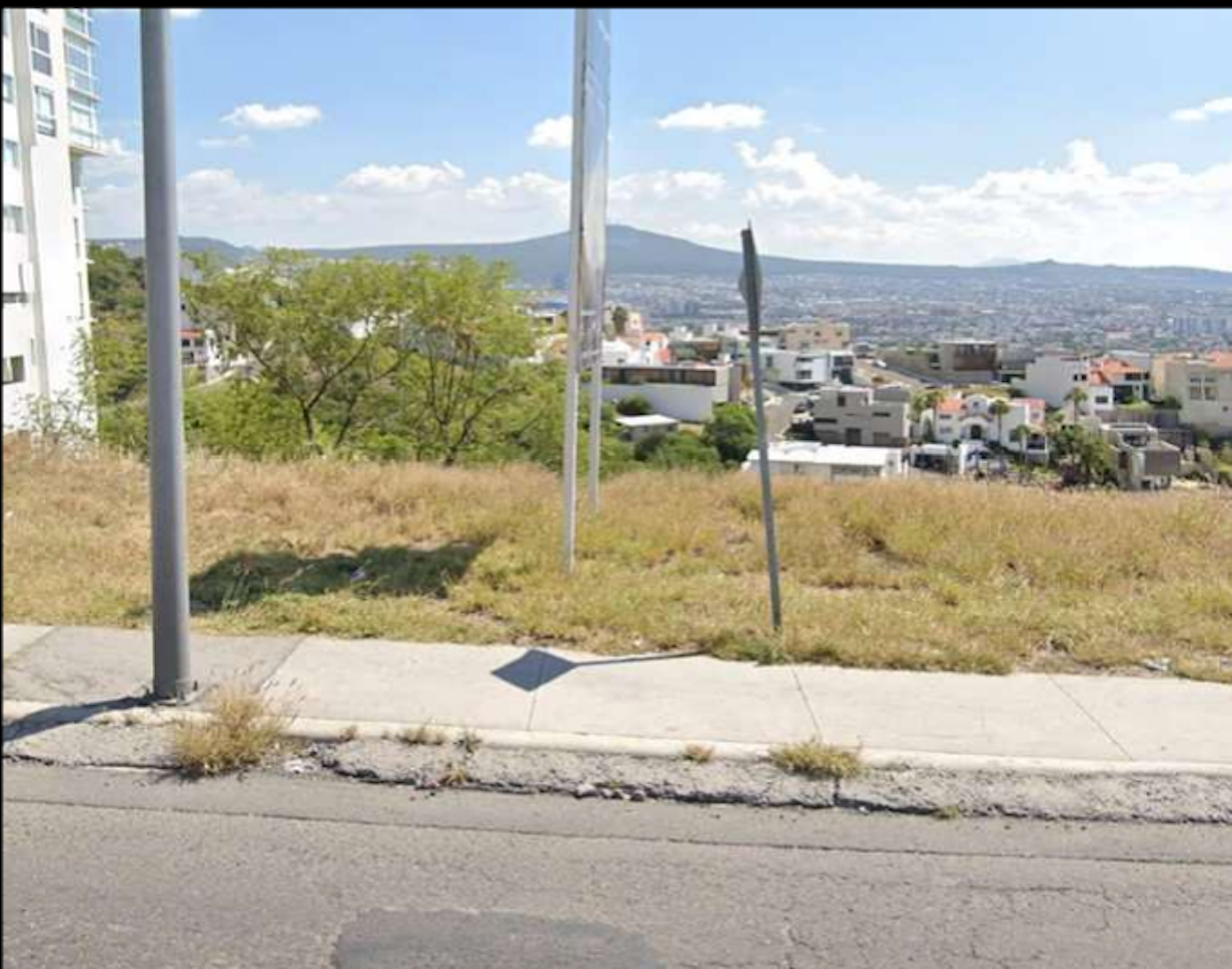 La imagen muestra una vista panorámica de una ciudad desde una zona elevada. Se observa un terreno baldío en primer plano con vegetación seca, seguido de una zona residencial con casas y edificios. Al fondo se aprecia una montaña y un cielo azul con nubes. La escena sugiere un área urbana en desarrollo con potencial para nuevos proyectos inmobiliarios, ofreciendo vistas atractivas de la ciudad y el paisaje circundante.