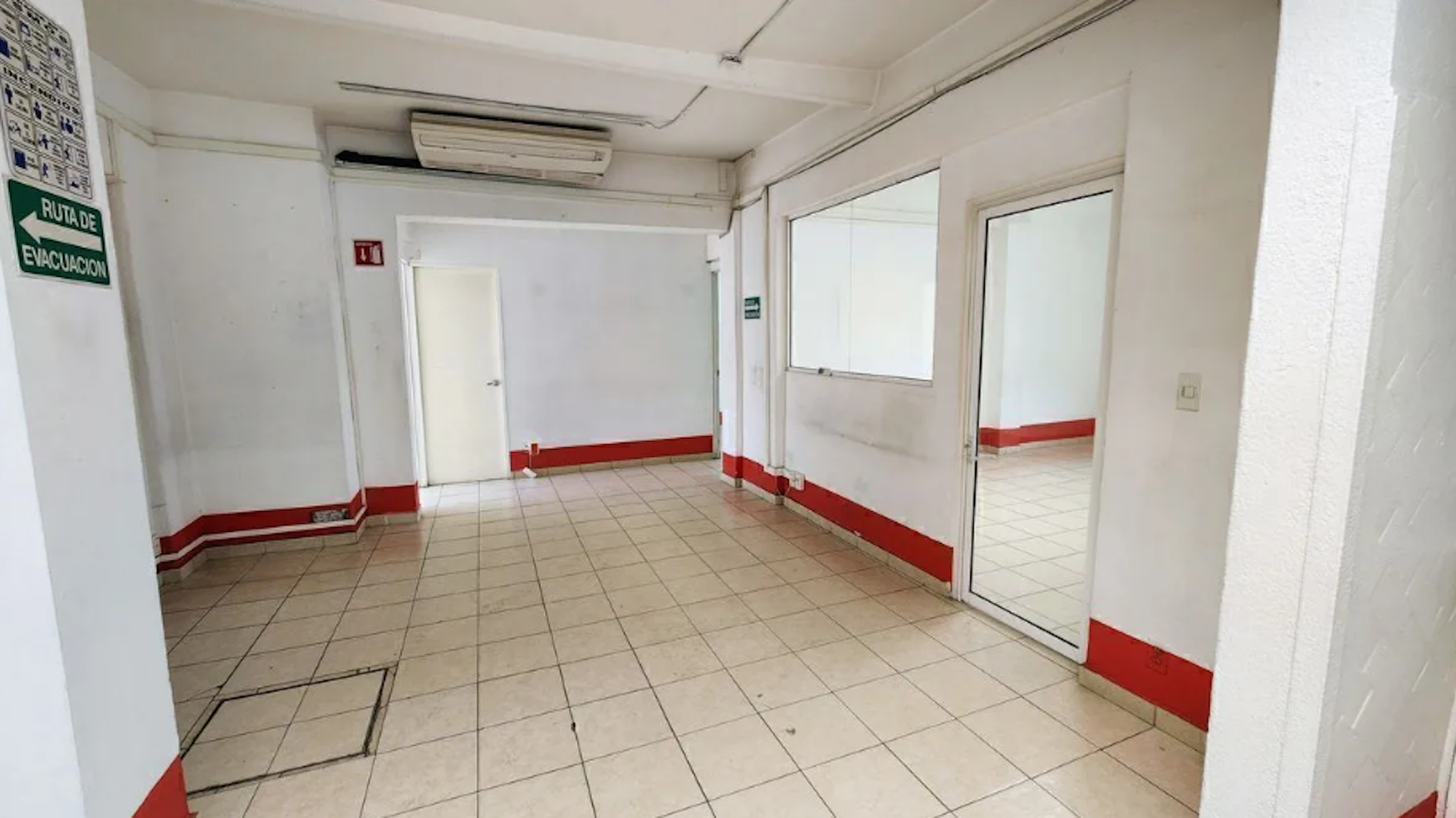 La imagen muestra un pasillo interior de un edificio, posiblemente una oficina o institución. Las paredes son blancas con un zócalo rojo, el piso es de baldosas beige. Se ven puertas blancas, ventanas interiores y un cartel de Ruta de evacuación. Hay un aire acondicionado en el techo. El espacio parece limpio pero algo austero, típico de un entorno institucional o comercial básico.