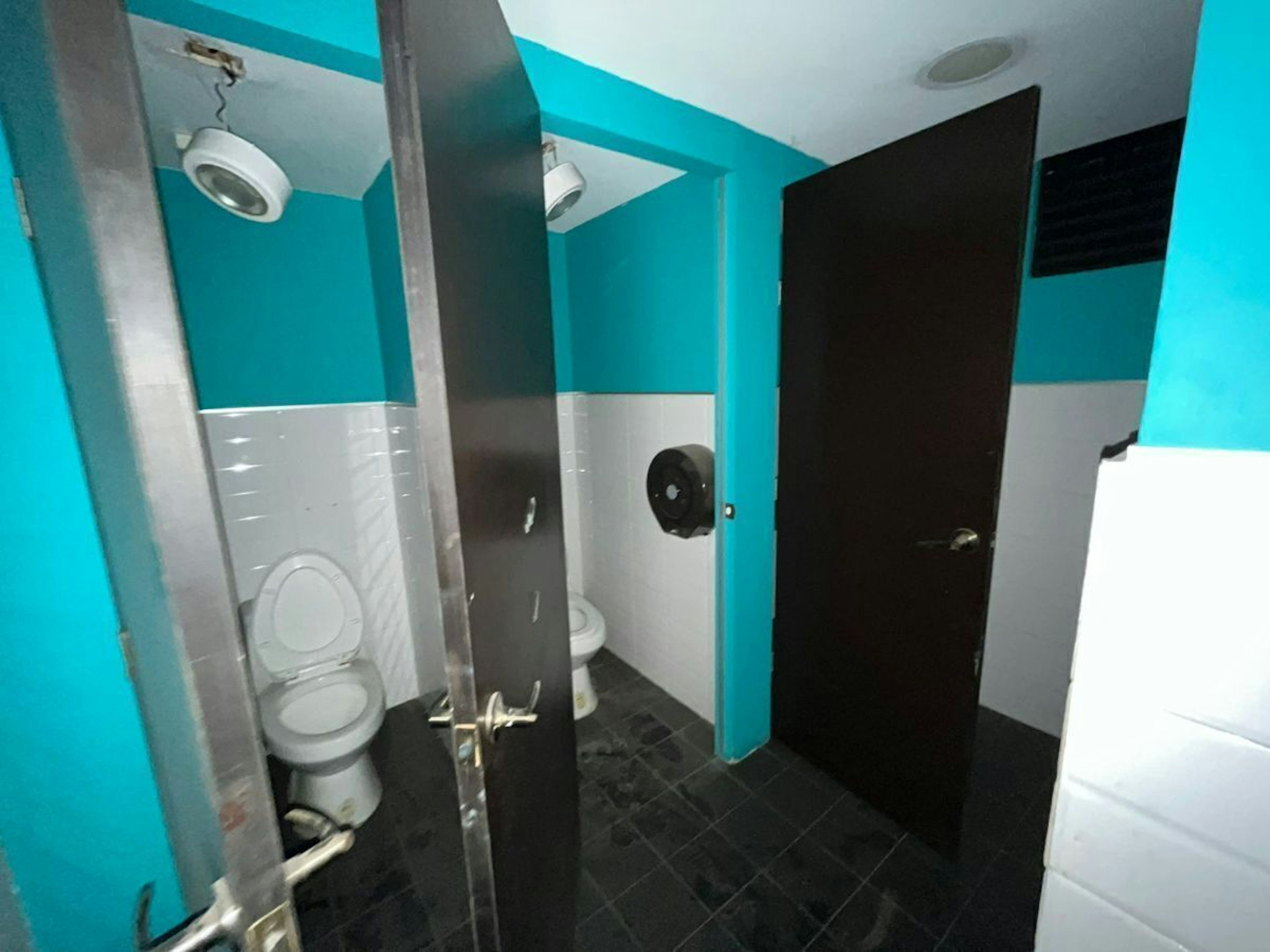 Imagen de un baño público con múltiples cabinas, mostrando inodoros y puertas parcialmente abiertas. Las paredes son de color azul y blanco, y el suelo es de baldosas oscuras.