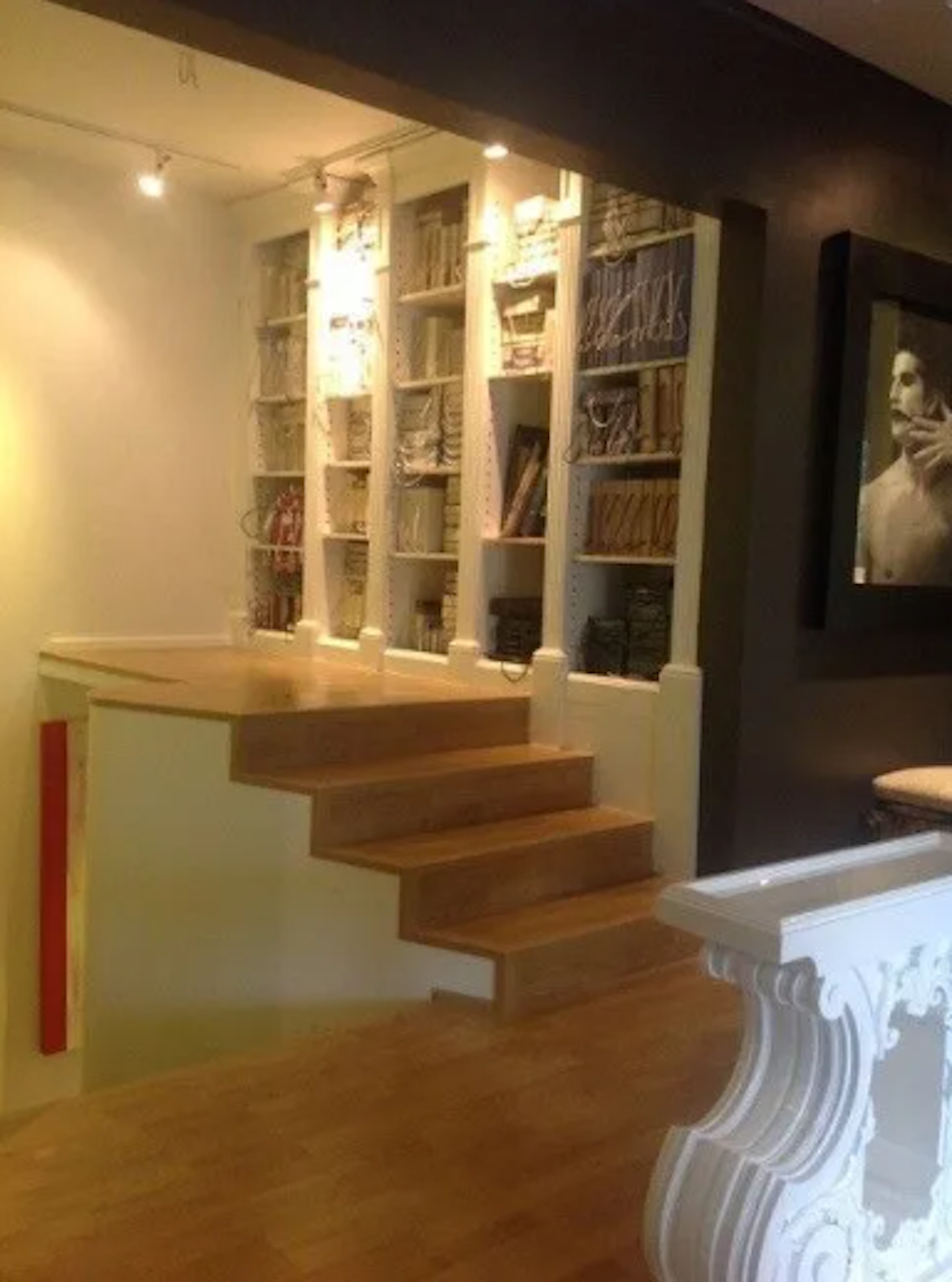 Esta imagen muestra un espacio interior elegante con una escalera de madera que conduce a una biblioteca empotrada iluminada. La biblioteca tiene estantes blancos llenos de libros y objetos decorativos. Junto a la escalera hay una ornamentada consola blanca. En la pared oscura se aprecia una obra de arte. El diseño combina elementos clásicos y modernos, creando un ambiente acogedor y sofisticado.