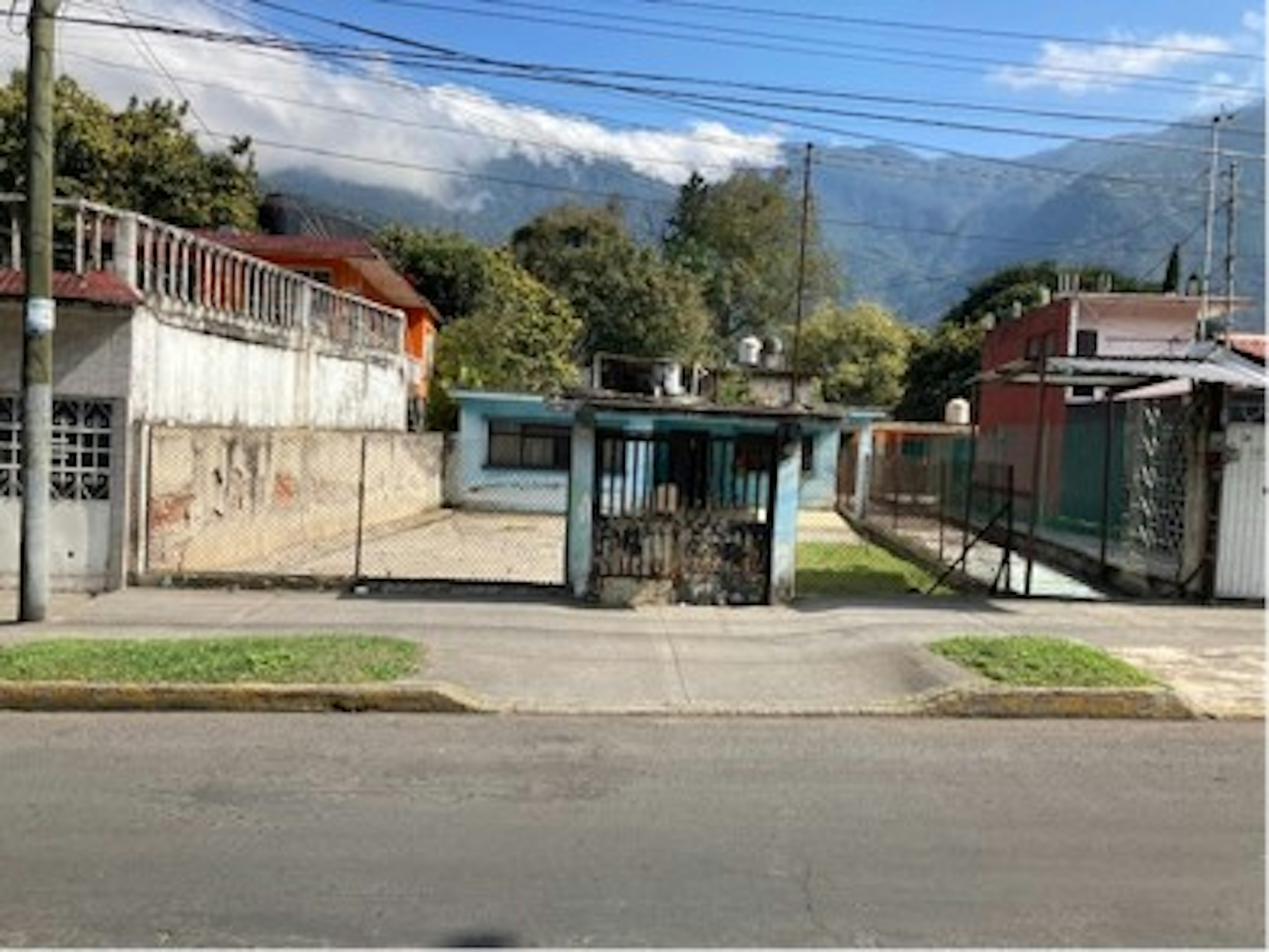 La imagen muestra una calle residencial en una zona montañosa. Se observan varias casas modestas de diferentes colores, con muros y rejas. Hay una pequeña estructura azul que parece ser una garita o caseta de seguridad. El entorno es rural o suburbano, con cables eléctricos visibles y montañas en el fondo. La calle está pavimentada y hay árboles alrededor de las casas, sugiriendo un vecindario tranquilo de clase media-baja.