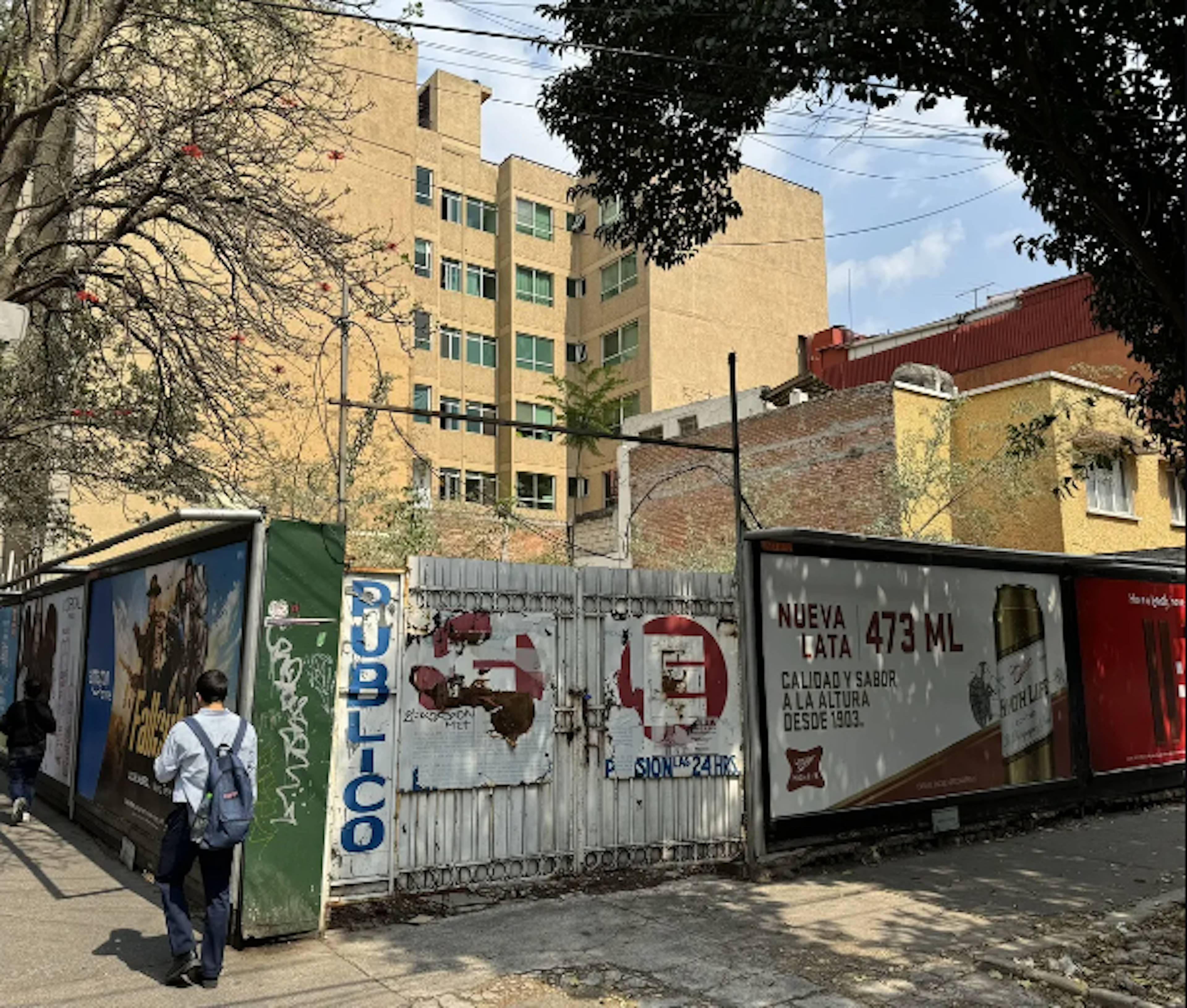 La imagen muestra un entorno urbano con un edificio de apartamentos de varios pisos en el fondo, de color beige. En primer plano hay una calle con vallas publicitarias y graffiti, incluyendo un anuncio de cerveza. Se observa un contraste entre la arquitectura moderna y elementos más antiguos o deteriorados, típico de áreas urbanas en desarrollo o transición. La escena sugiere una zona residencial y comercial mixta.