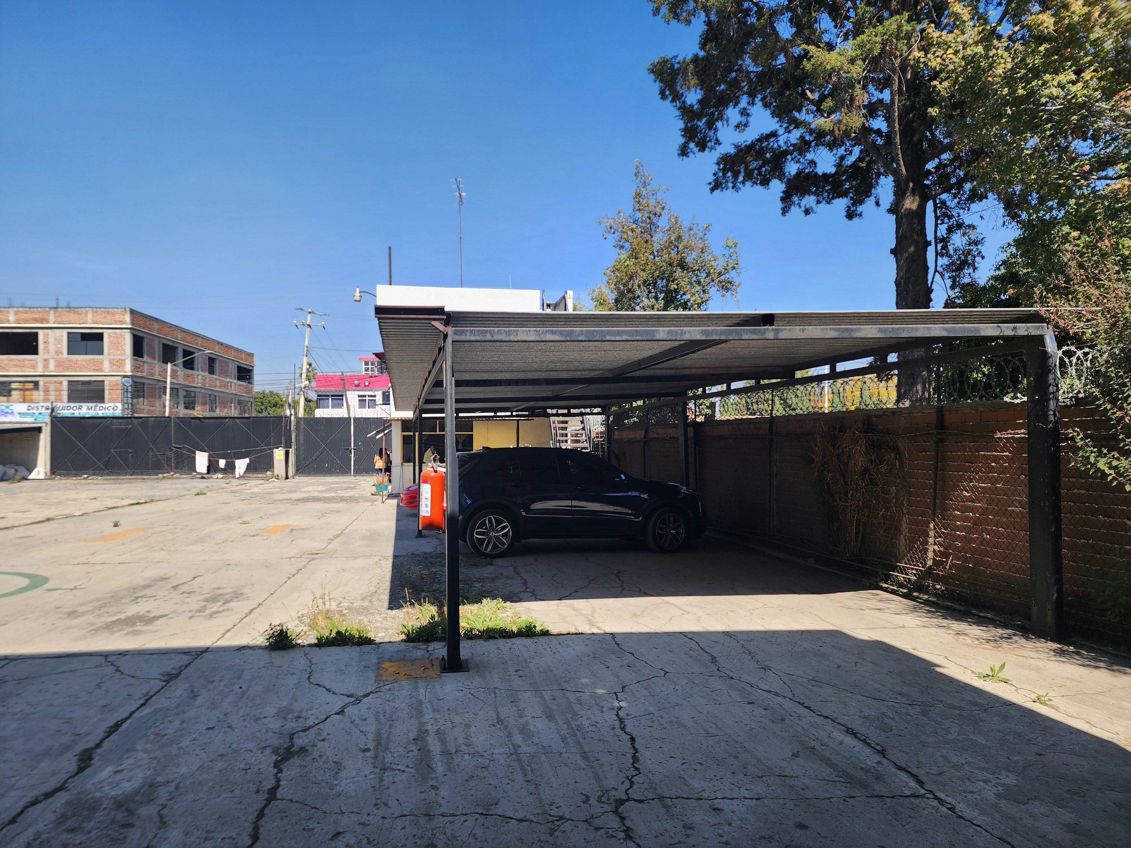 La imagen muestra un estacionamiento techado en un entorno urbano. Se ve un vehículo negro estacionado bajo una estructura metálica que sirve como techo. El área circundante incluye un patio de concreto agrietado, un edificio de ladrillo en construcción al fondo, y algunos árboles. El cielo es azul claro, lo que sugiere un día soleado. Este tipo de espacio podría ser parte de una propiedad residencial o comercial, ofreciendo estacionamiento cubierto.