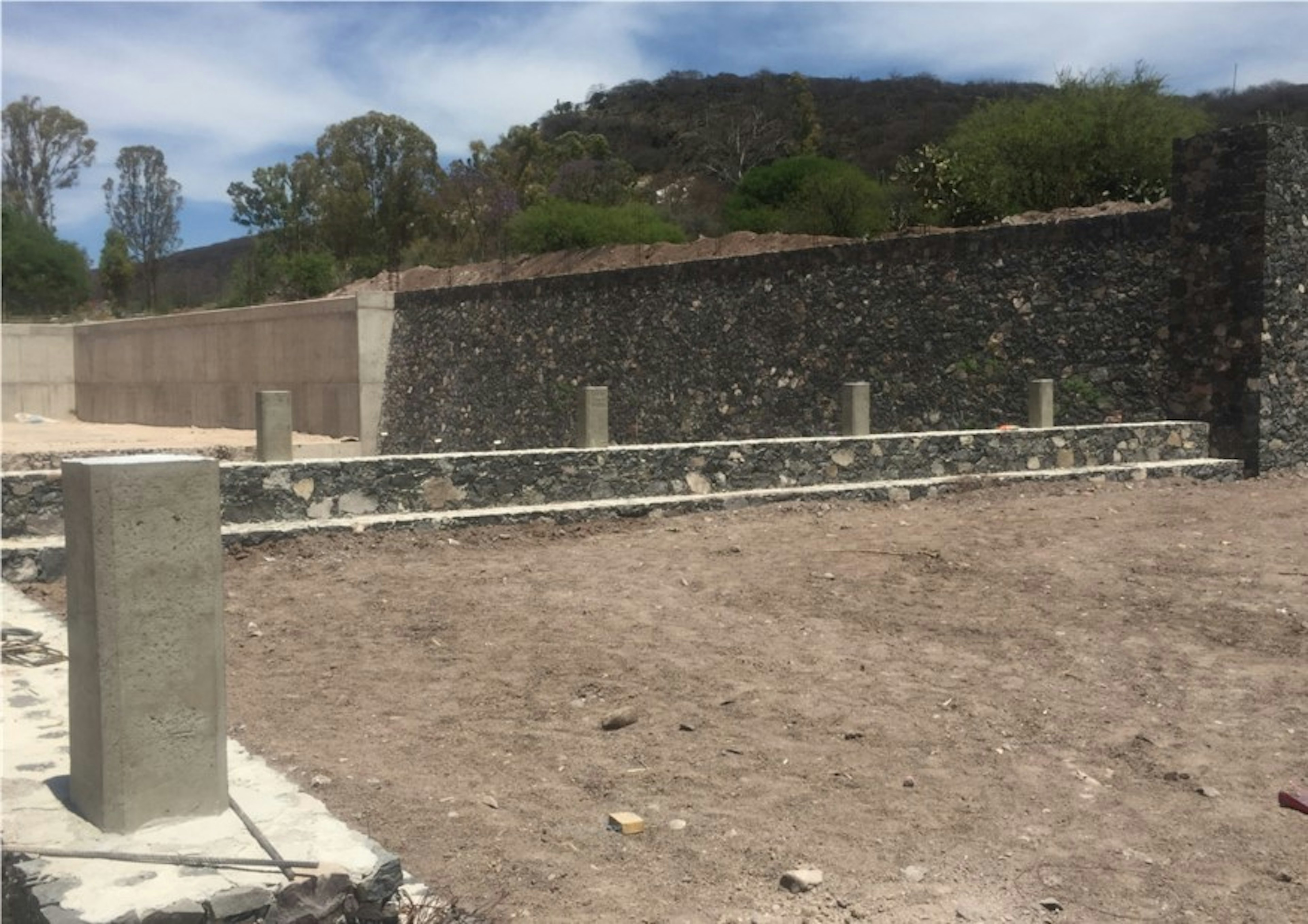 La imagen muestra un terreno en construcción con cimientos y muros de piedra parcialmente erigidos. El cielo es claro y hay vegetación al fondo. Ideal para desarrollo inmobiliario.